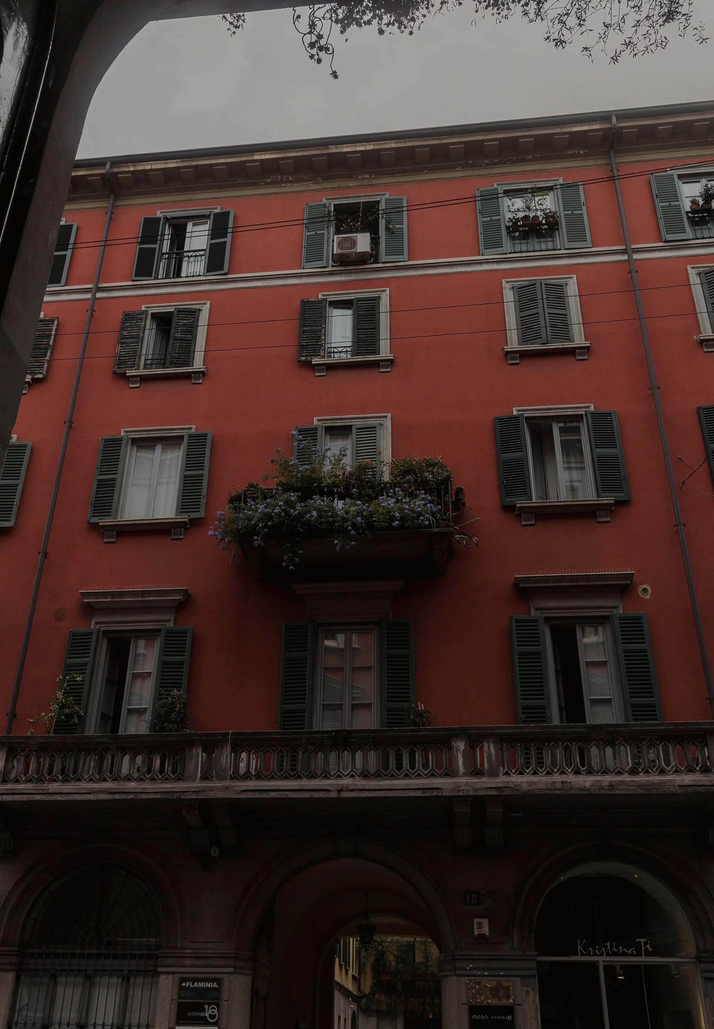 Facciata di un palazzo milanese color mattone con persiane verdi e balconi in ferro, vista dal basso