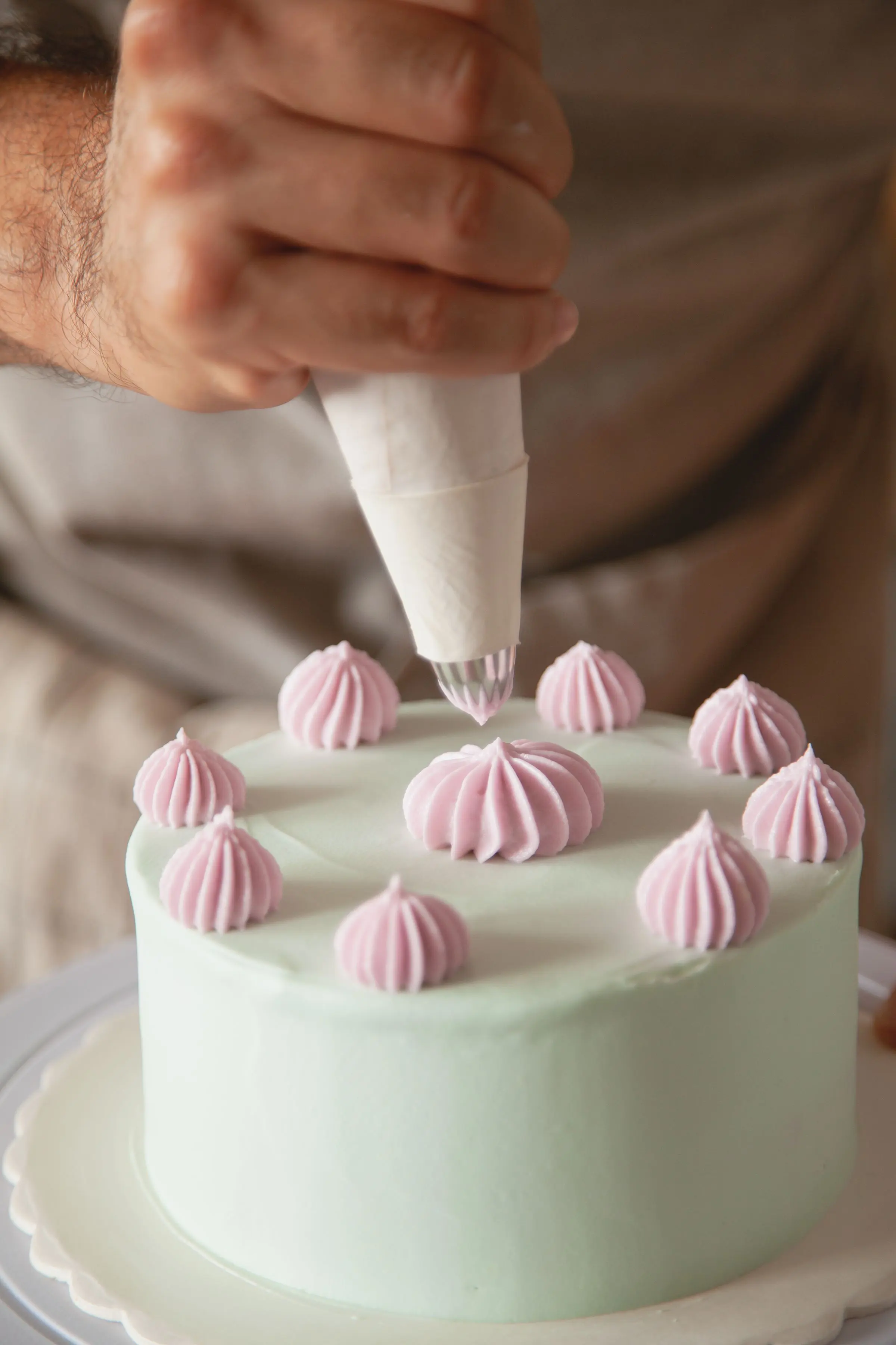 Una mano spreme meringhe rosa con la sac à poche sopra una torta verde pistacchio