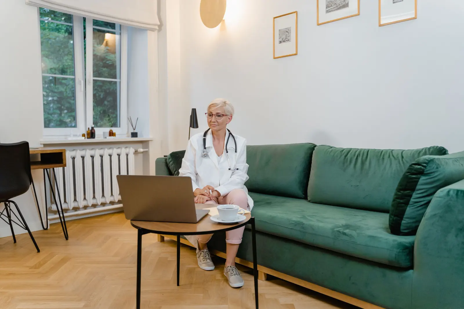 Una medica in camice segue un corso dal portatile, seduta sul divano di casa