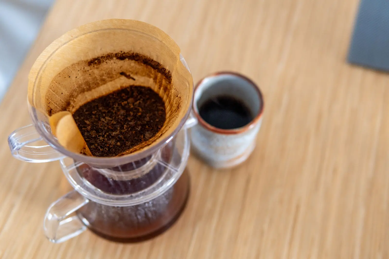 Un filtro di vetro con il fondo di caffè e una tazzina su un tavolo di legno chiaro