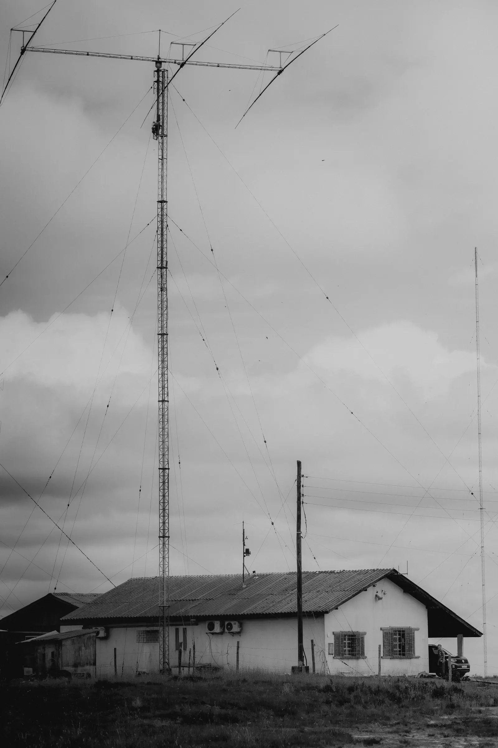 Antenne filari e un traliccio accanto a un edificio basso in aperta campagna, in bianco e nero