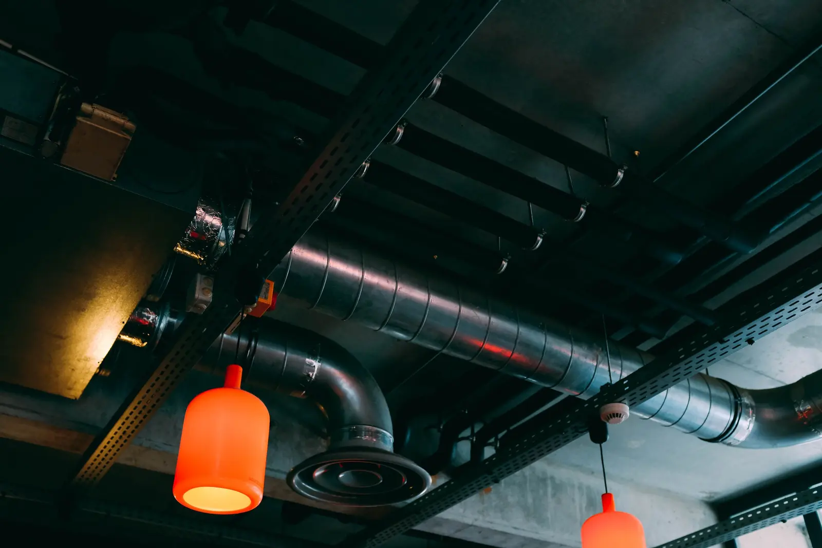 Soffitto scuro di un locale con i canali dell'aria a vista, un diffusore circolare e lampade arancioni