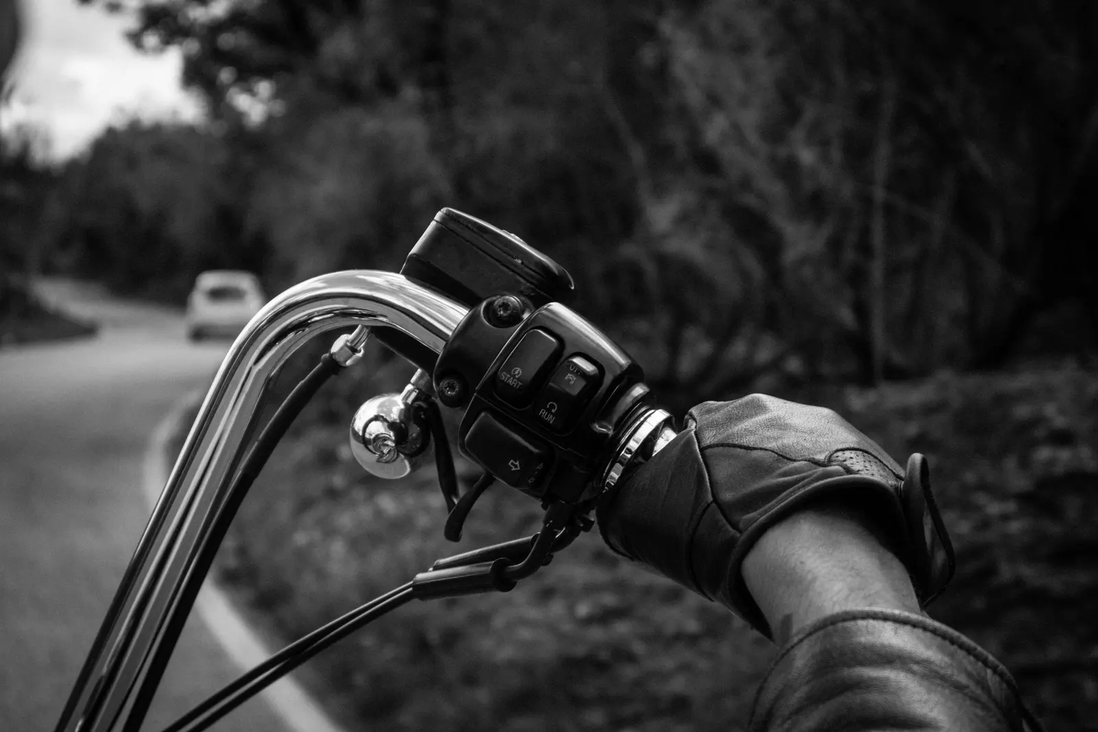 Mano con guanto di pelle sul manubrio di una moto, la strada che curva davanti, in bianco e nero