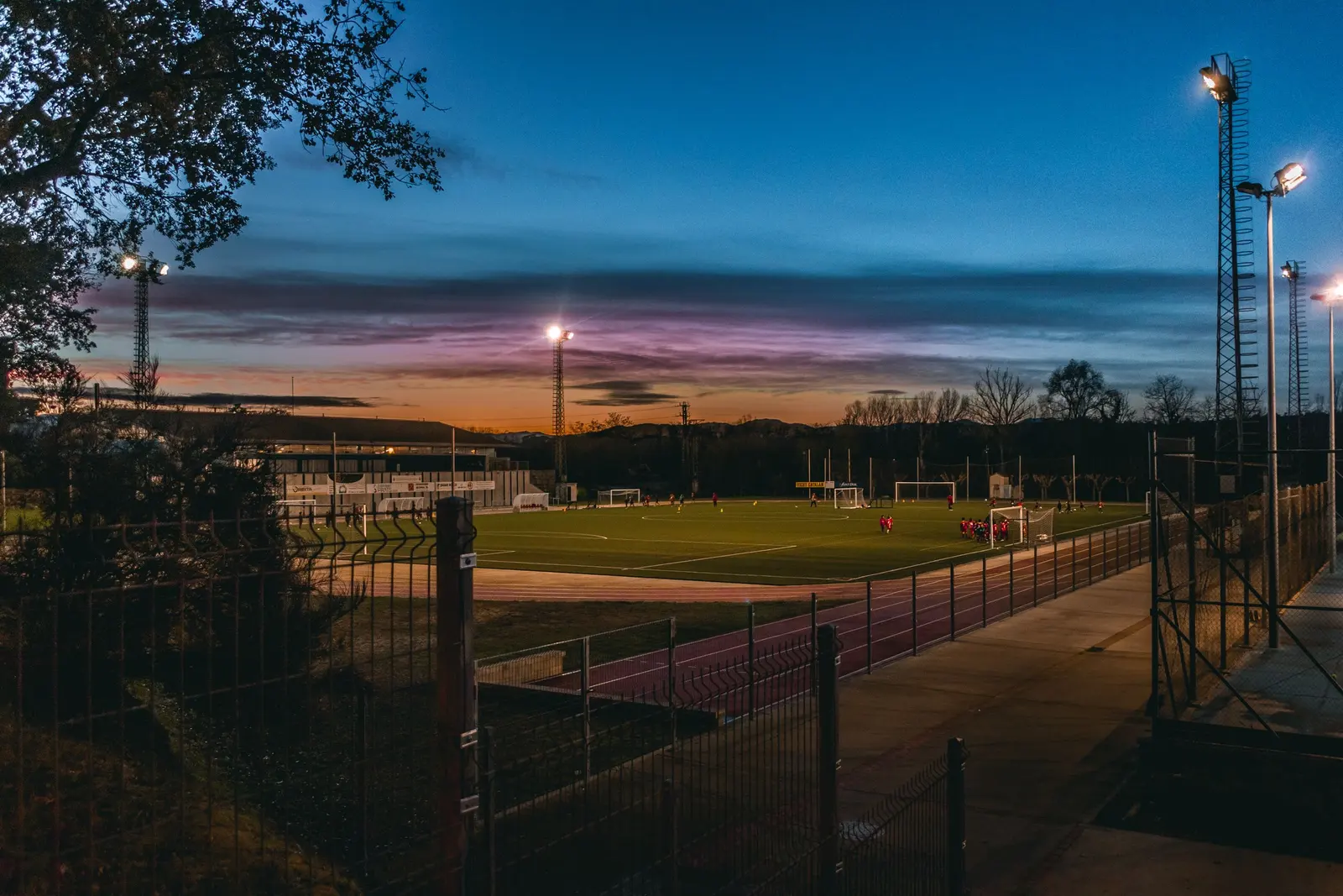 Il centro sportivo al tramonto, con le torri faro accese e la recinzione in primo piano