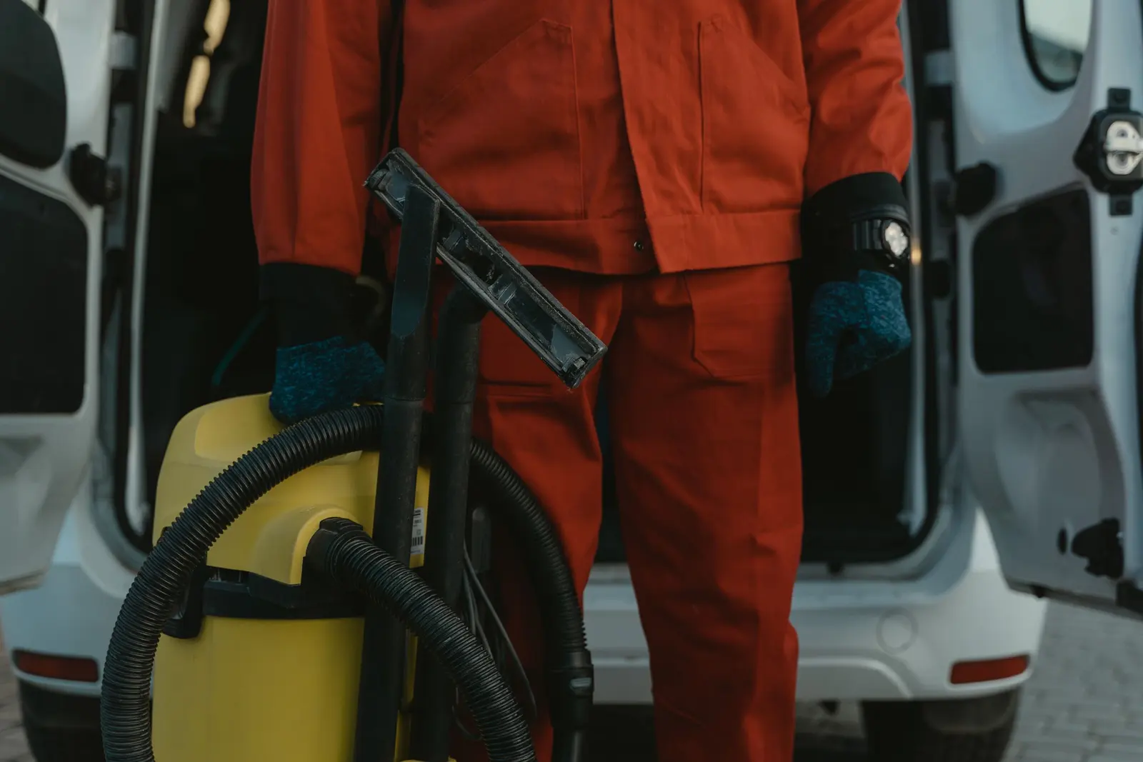 Un addetto in tuta arancione con un aspiratore giallo davanti al portellone aperto di un furgone