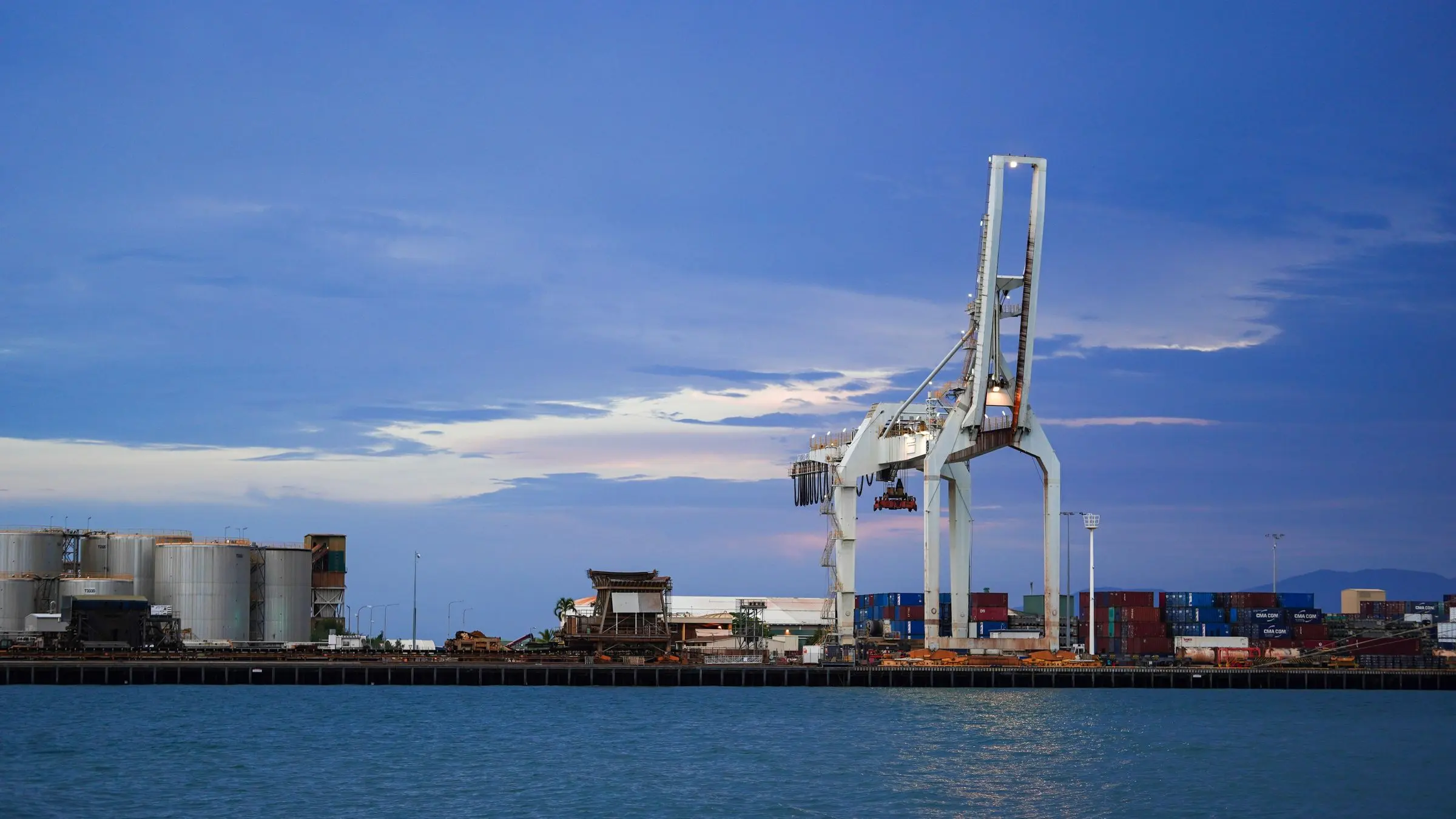 Una gru a cavalletto di un terminal container all'imbrunire, con i serbatoi sulla banchina e l'acqua del canale