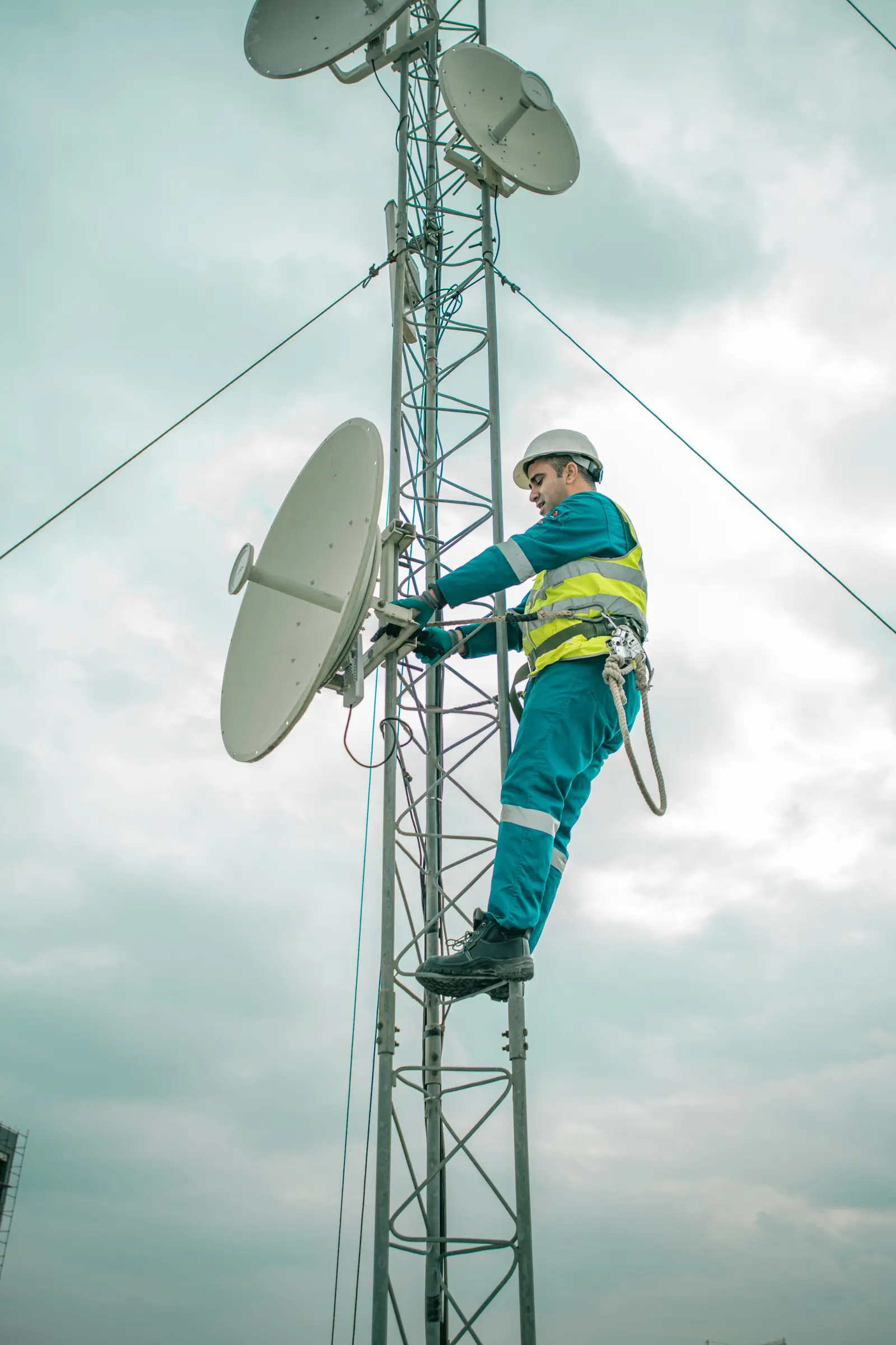 Un tecnico con casco e imbrago, agganciato a un traliccio, regola il supporto di un'antenna a parabola