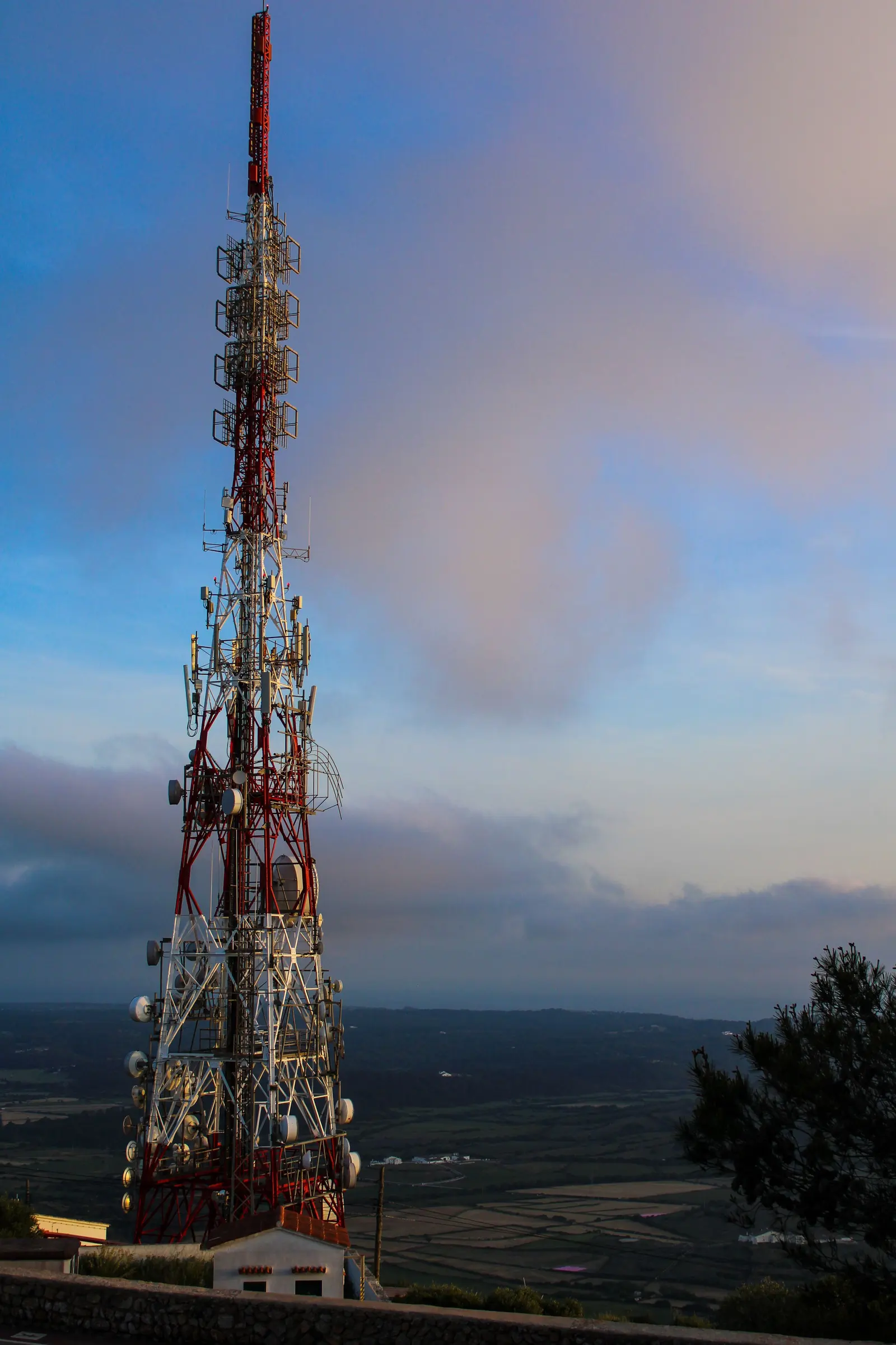 Un traliccio bianco e rosso carico di antenne, su un'altura che domina la pianura al tramonto