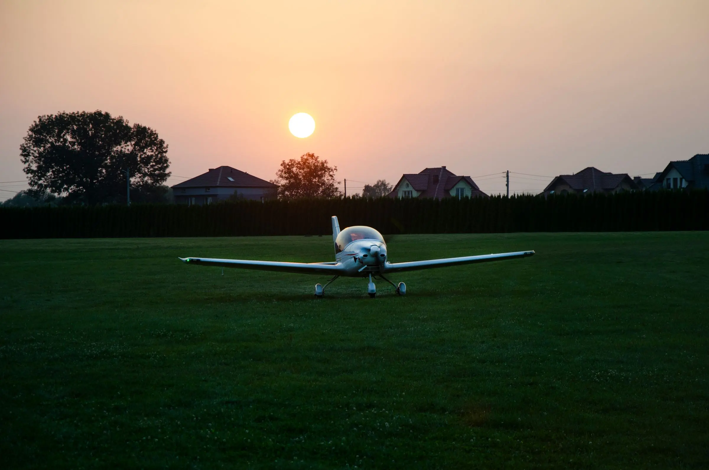 Un aereo leggero fermo sul prato del campo di volo al tramonto, con il sole basso dietro gli alberi
