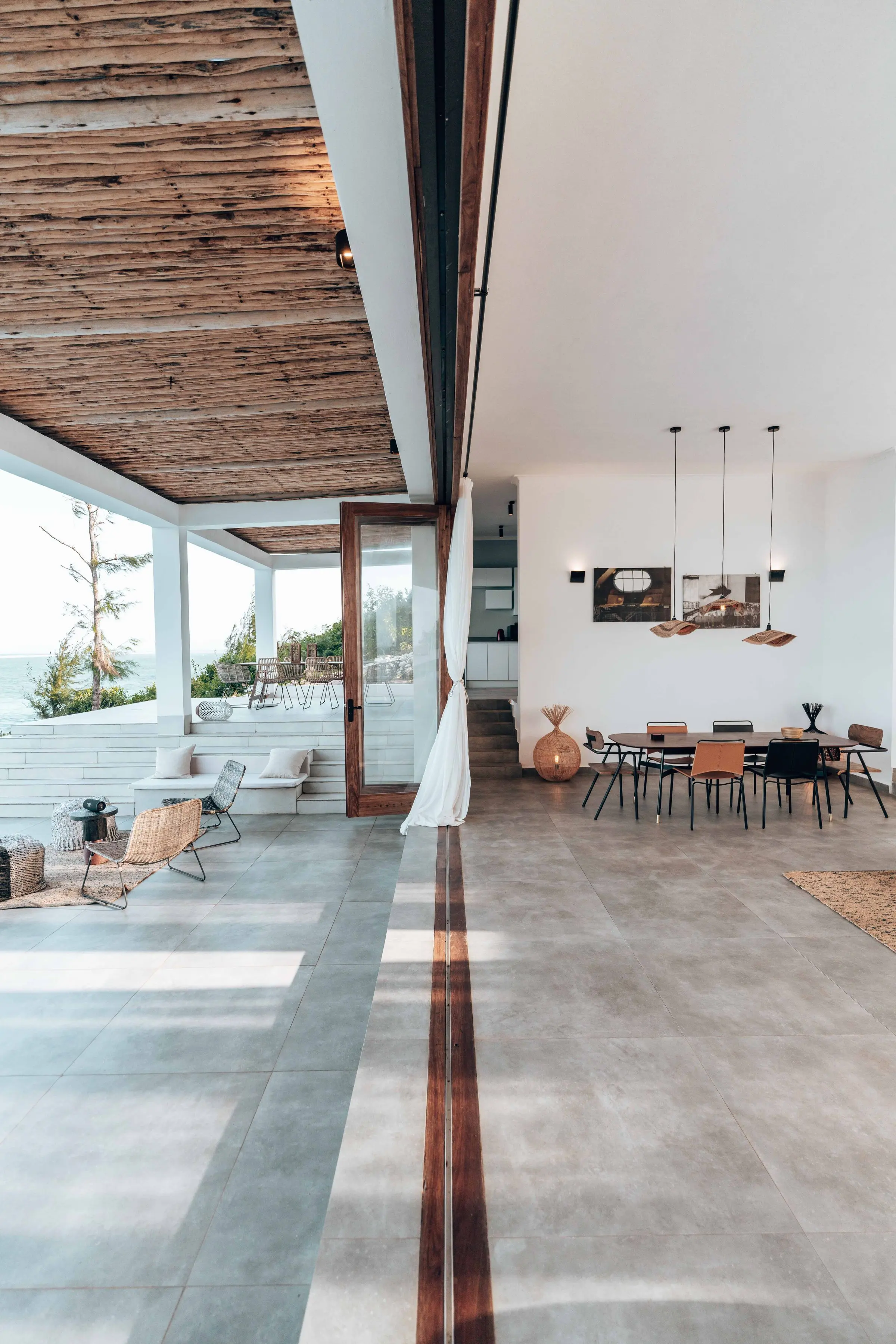 Il portico con il soffitto in canne si apre sul soggiorno bianco: stesso pavimento, tavolo da pranzo e lampade sospese