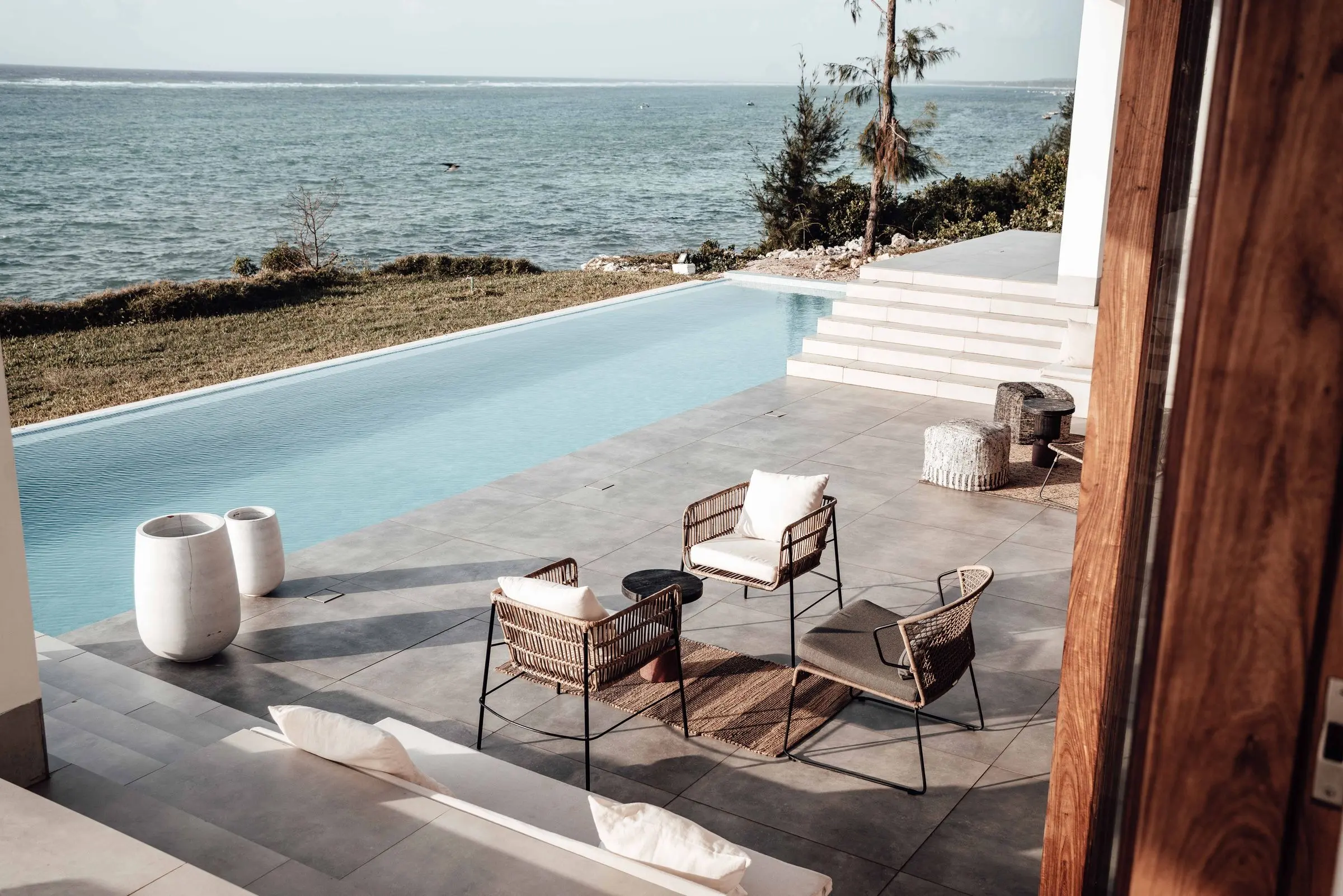Terrazza con sedute in corda accanto alla piscina, un prato e il mare aperto, vista dalla porta del soggiorno