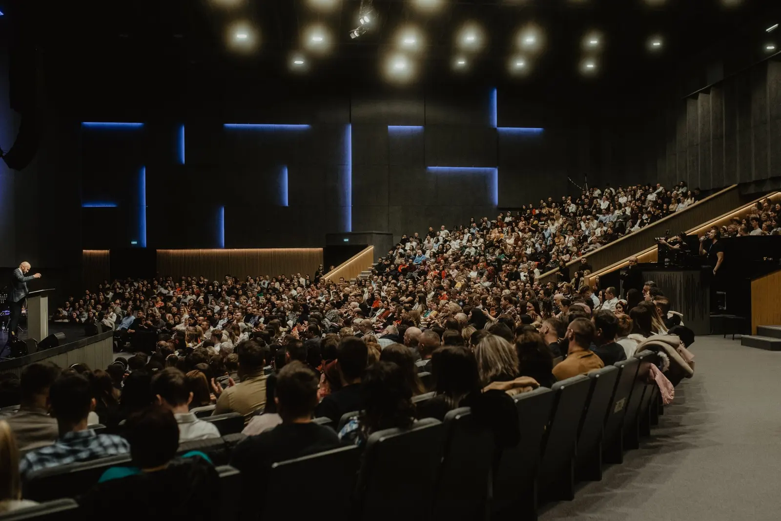 Un auditorium pieno con la platea in salita e un relatore al leggio, pareti scure con tagli di luce blu
