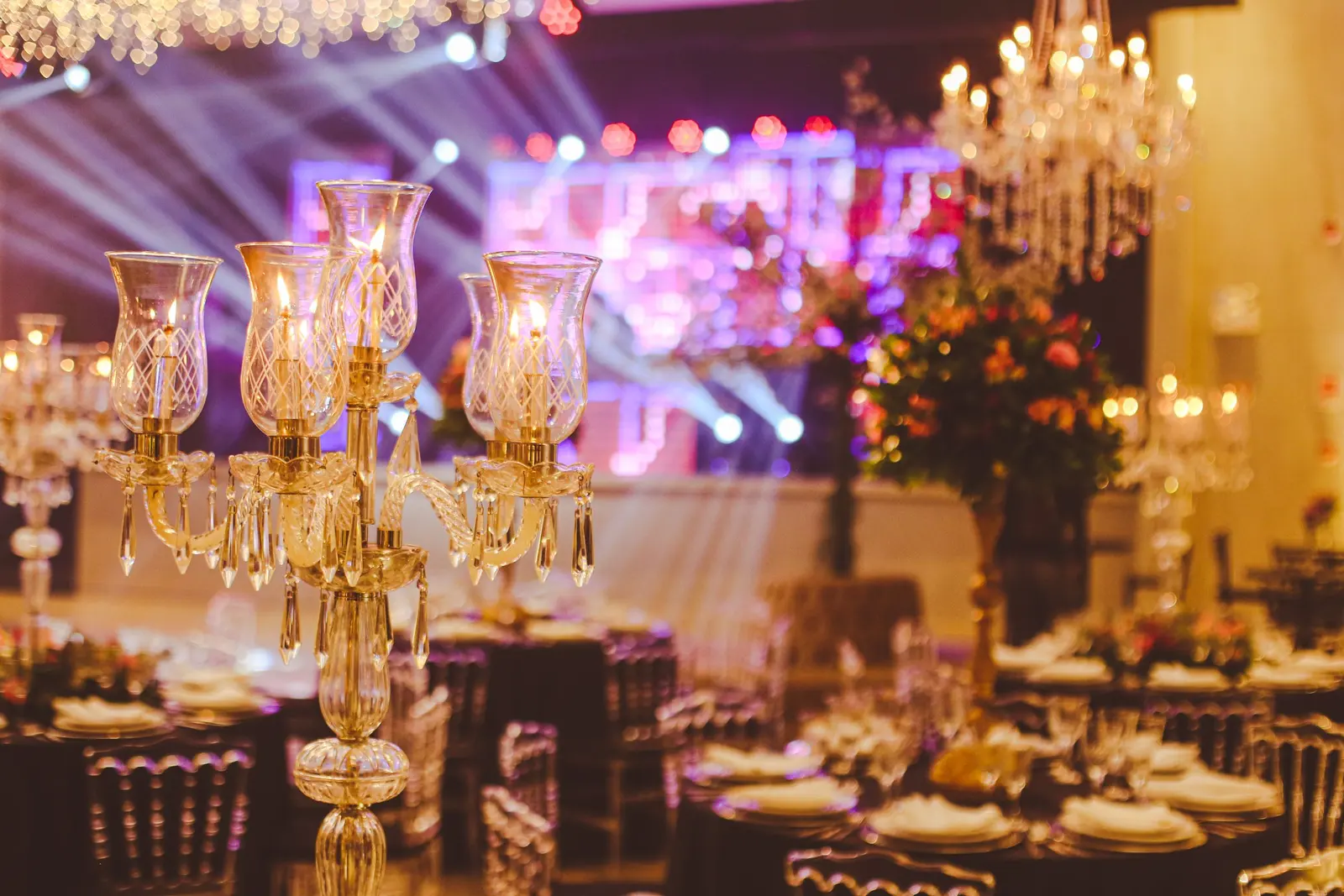 Sala di una cena di gala: candelabro di cristallo con candele accese in primo piano, tavoli apparecchiati e luci di scena dietro