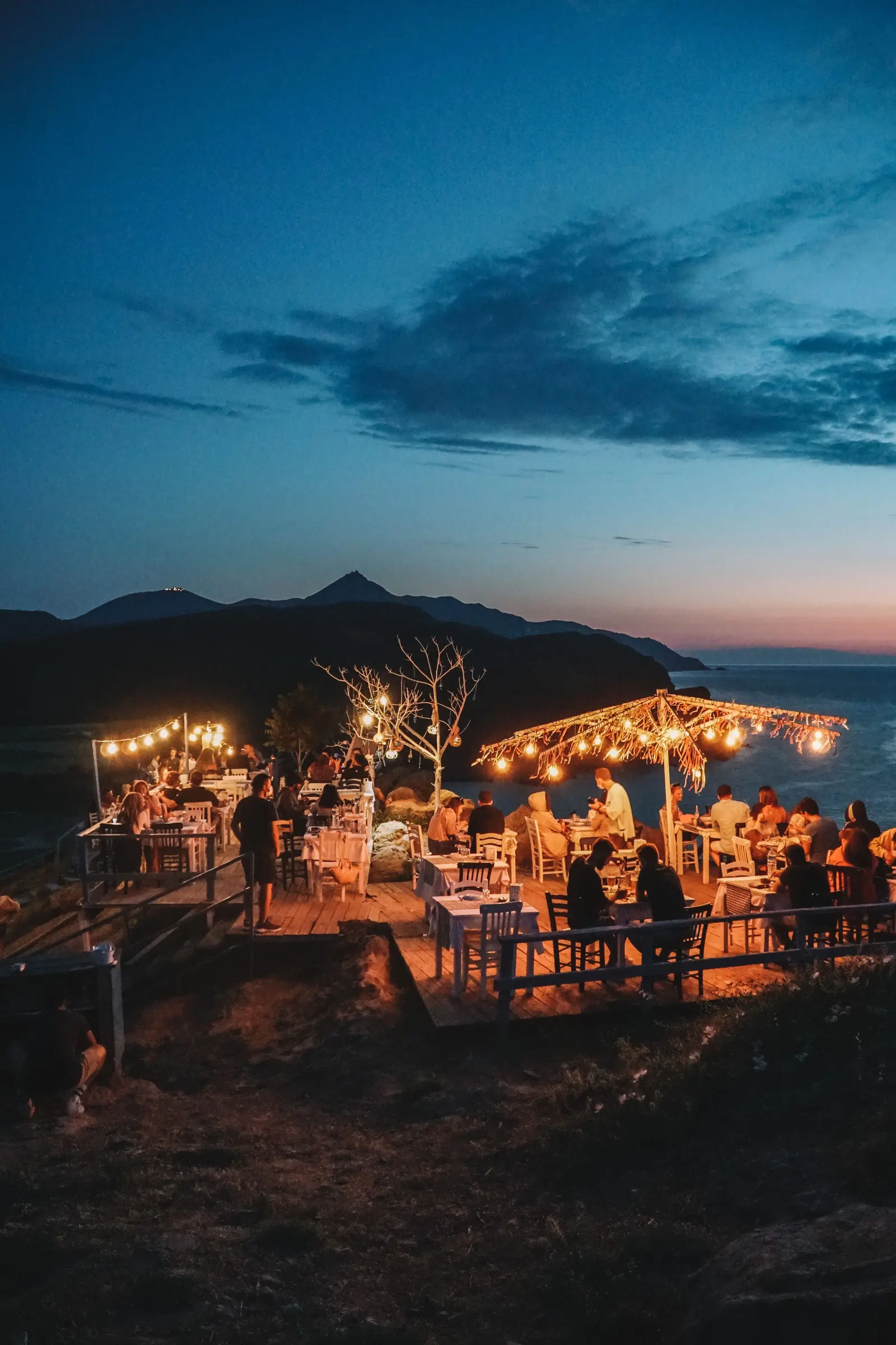 Terrazza di un ristorante sul mare all'imbrunire: tavoli pieni, luci appese a un pergolato, monti e cielo blu