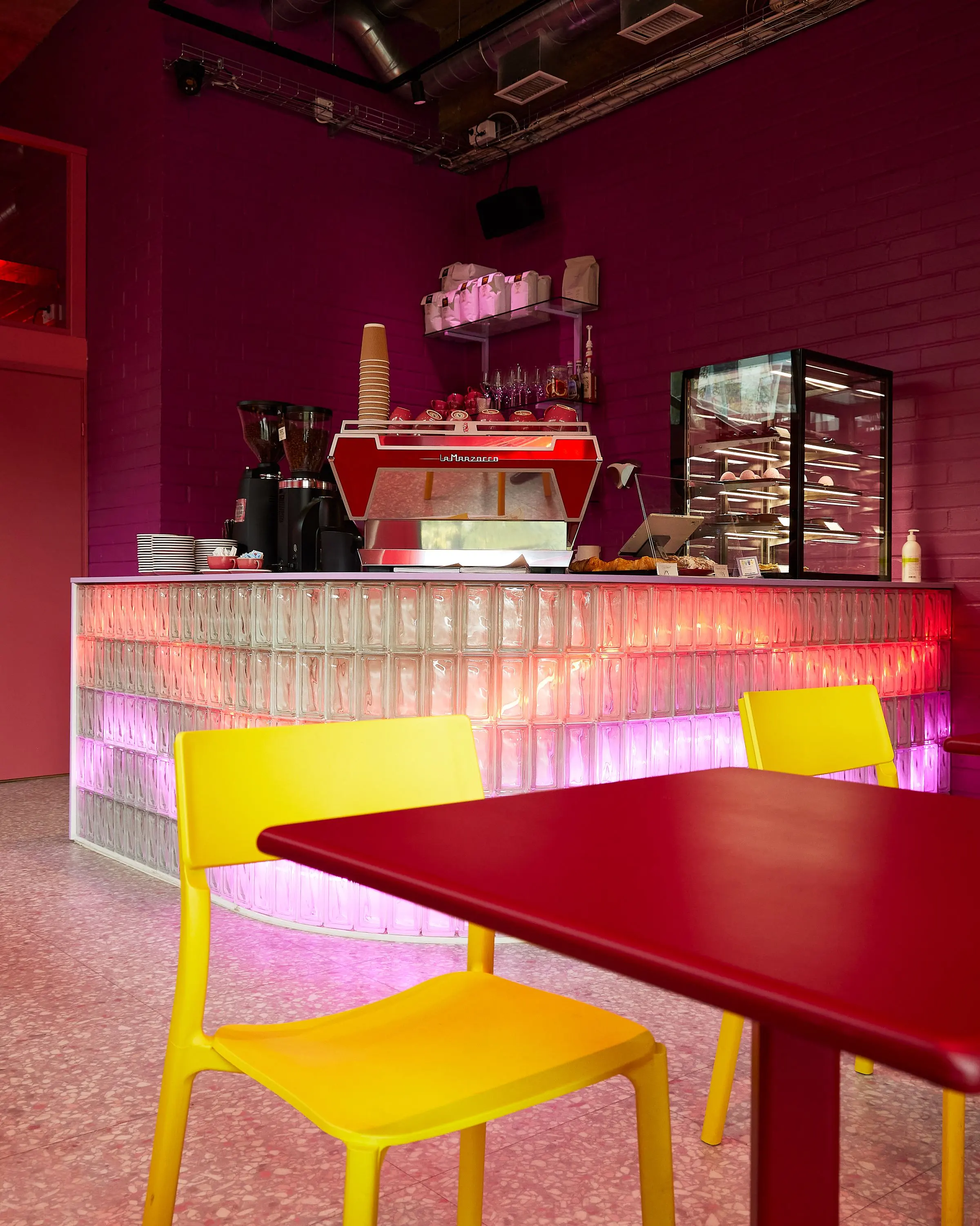 Bar con pareti magenta, banco in vetrocemento illuminato dall'interno, sedie gialle e tavolo rosso