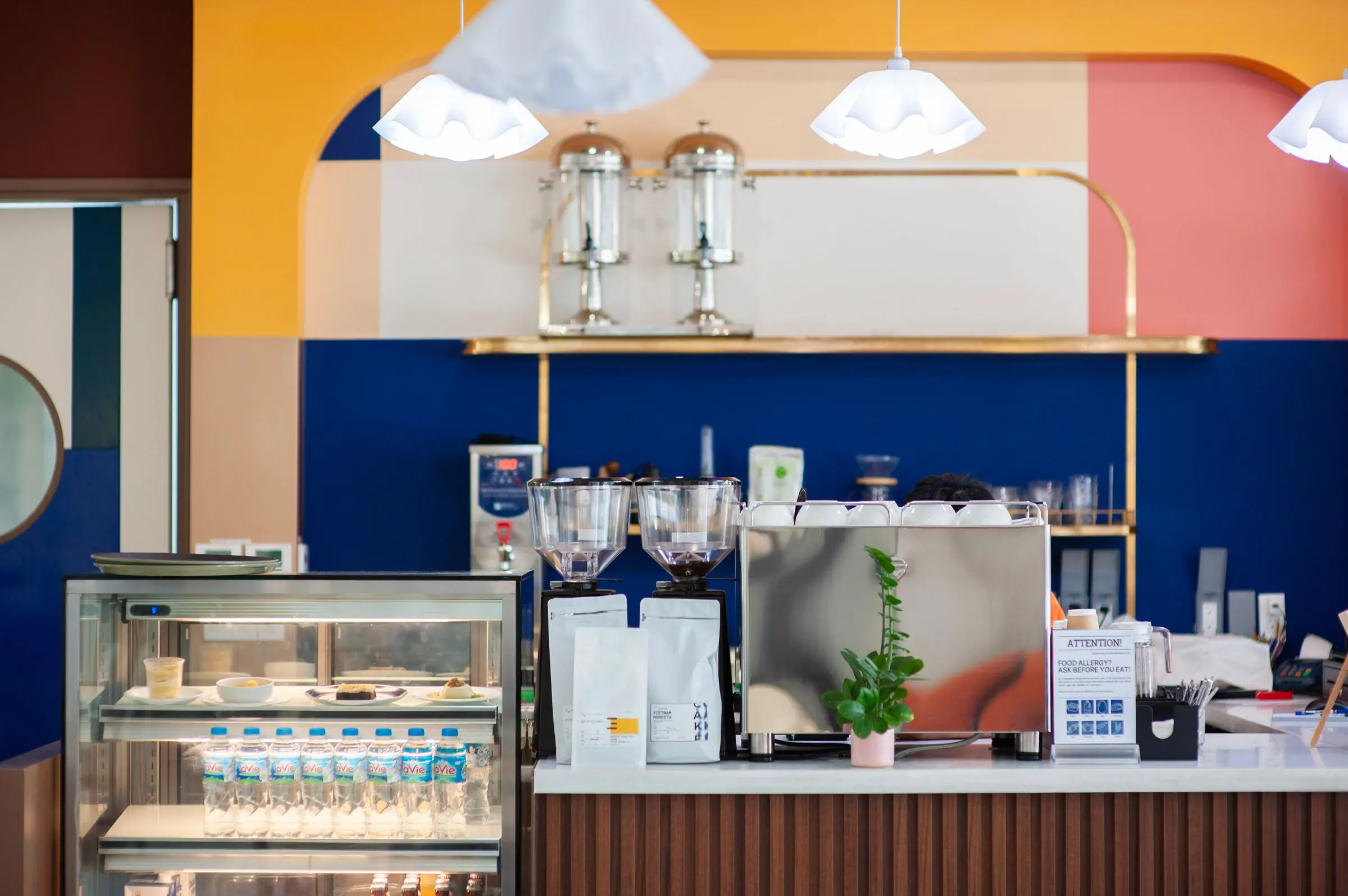 Banco di caffetteria con pareti a fasce gialla, rosa e blu, macchina del caffè e vetrina dei dolci