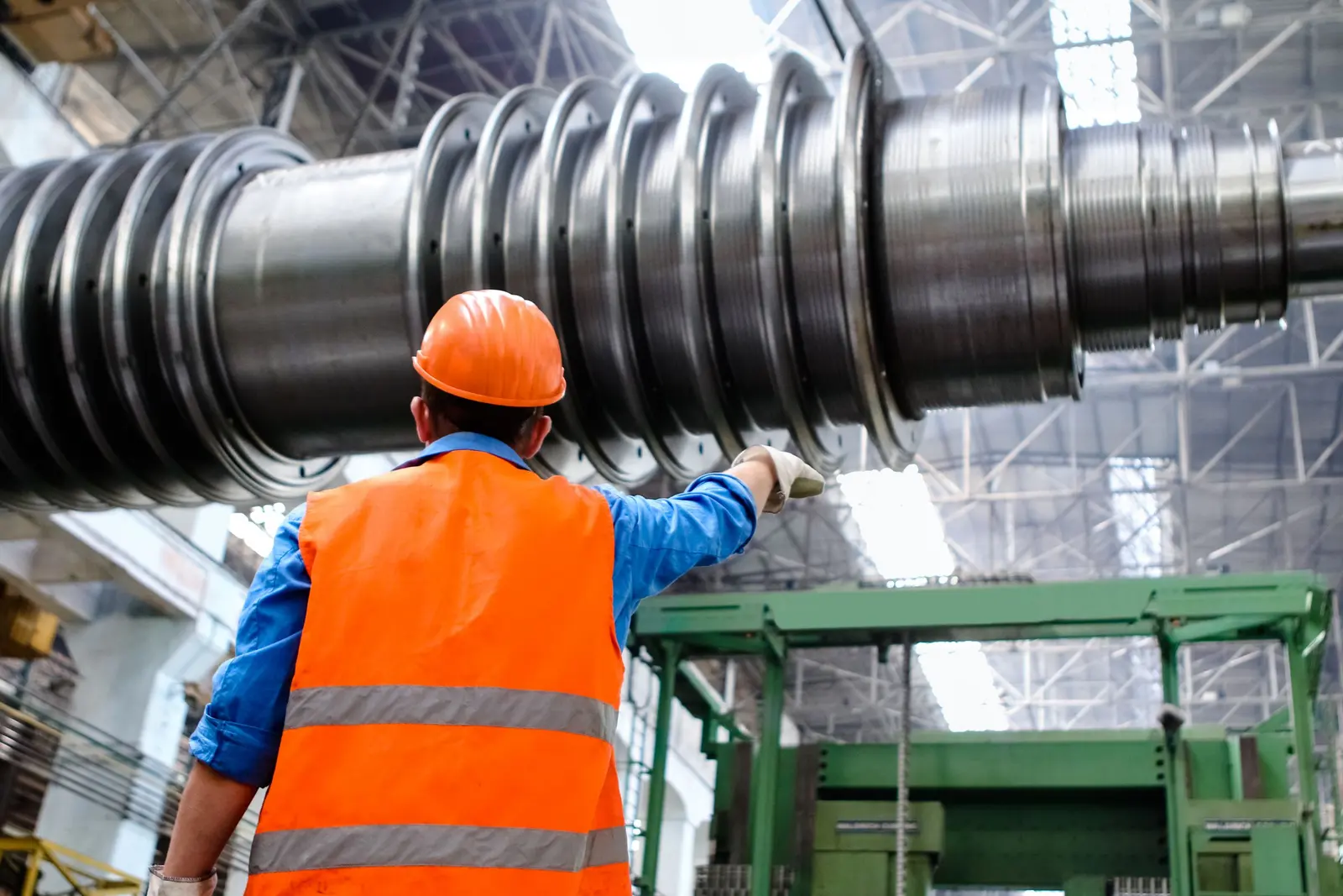 Un tecnico di spalle con elmetto e gilet arancione controlla un grande albero meccanico in officina