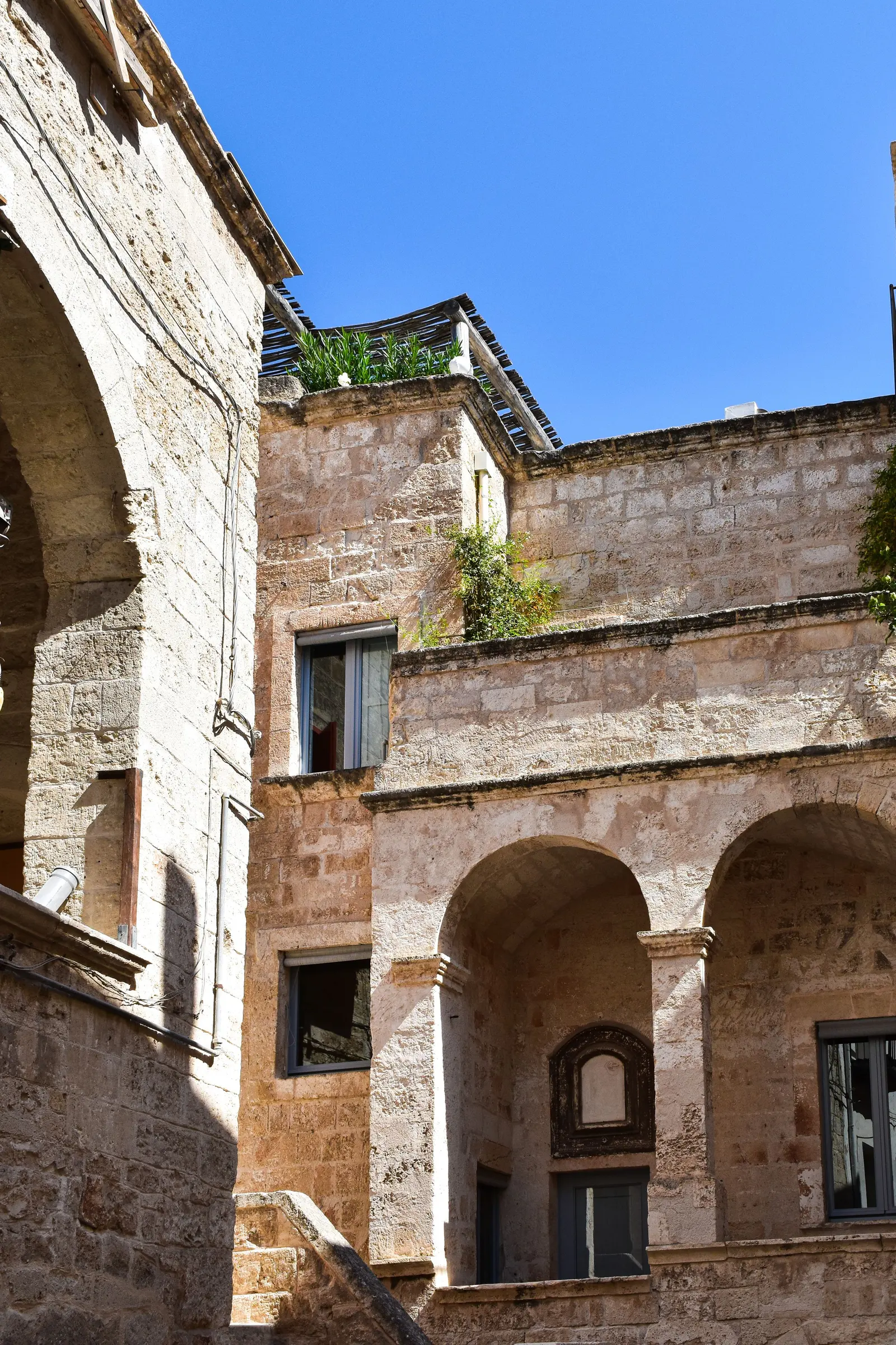 Cortile in pietra chiara con due arcate su colonne, una scala e una grande arcata a sinistra, sotto il cielo blu