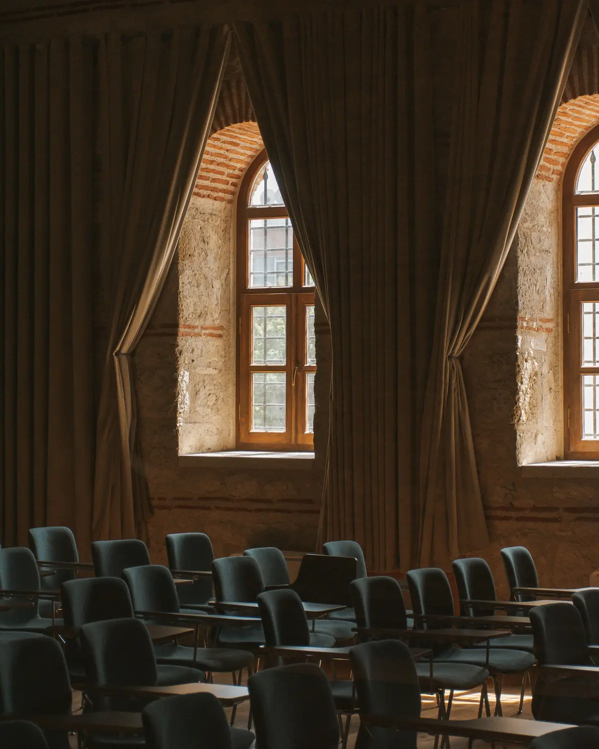 Sala con muri in pietra e mattoni, due finestre ad arco con tende scure e file di sedie imbottite con ribaltina