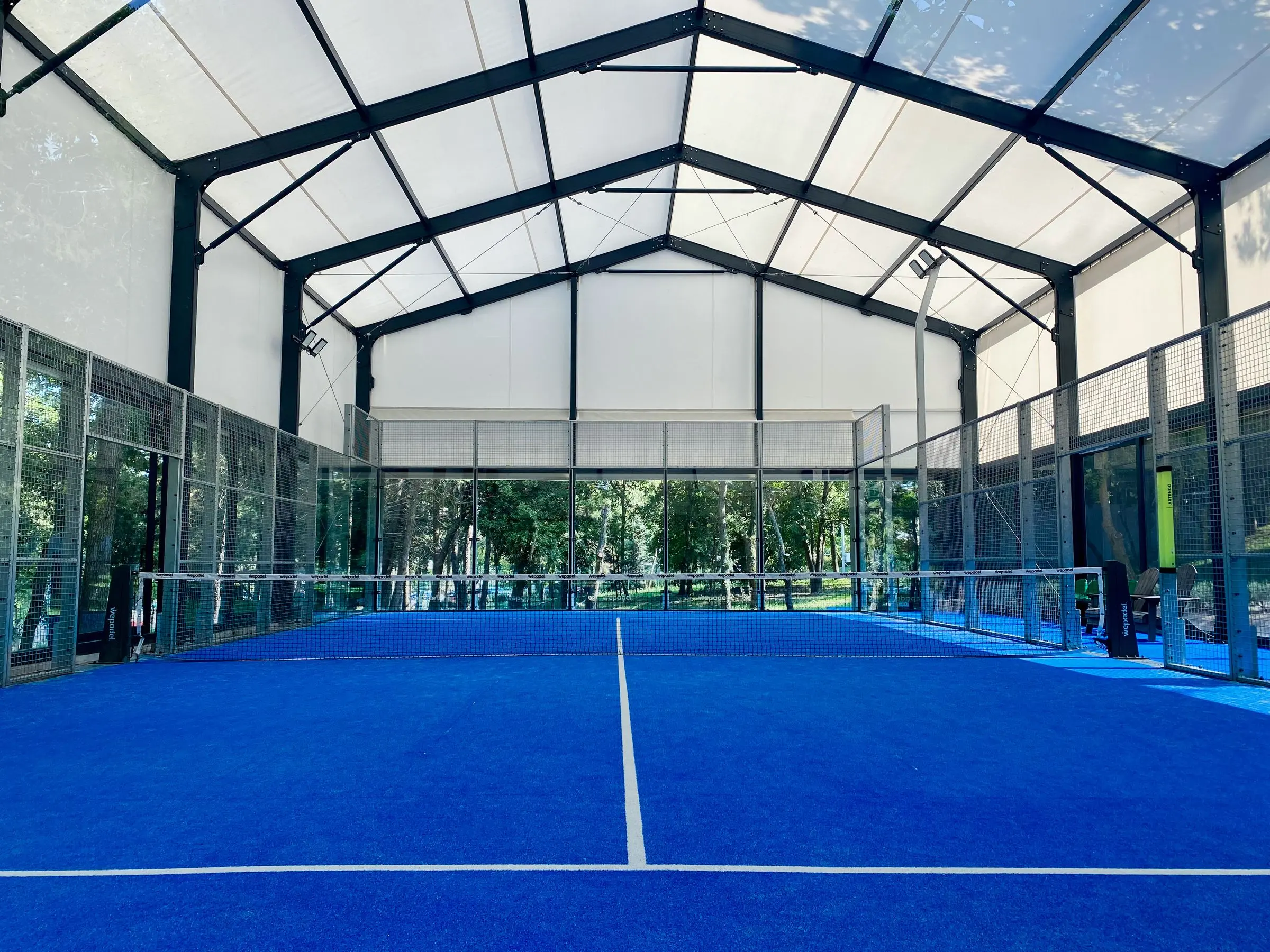 Campo da padel con il manto blu sotto una copertura a telo bianco, vetrate sui fondi e alberi all'esterno