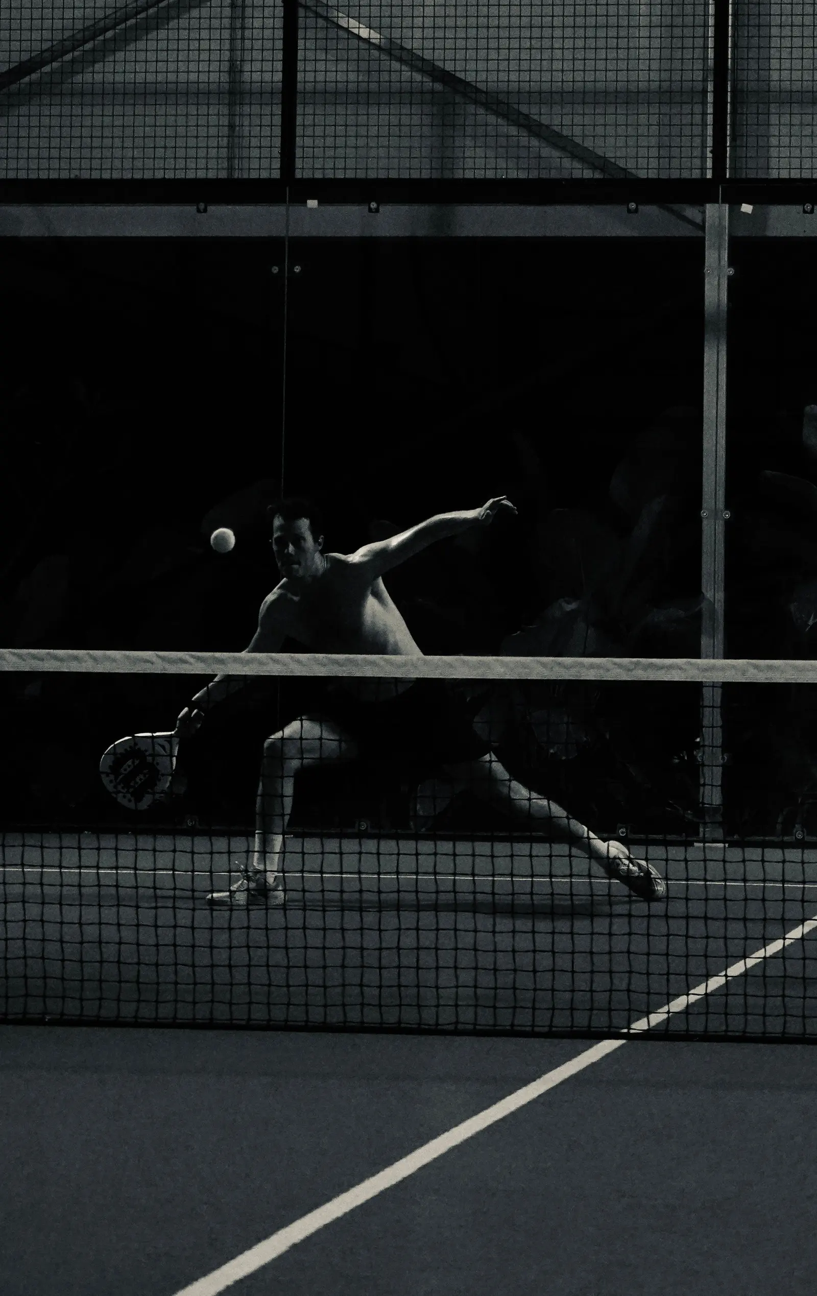 Un giocatore di padel colpisce in allungo sotto la rete, di sera, in bianco e nero