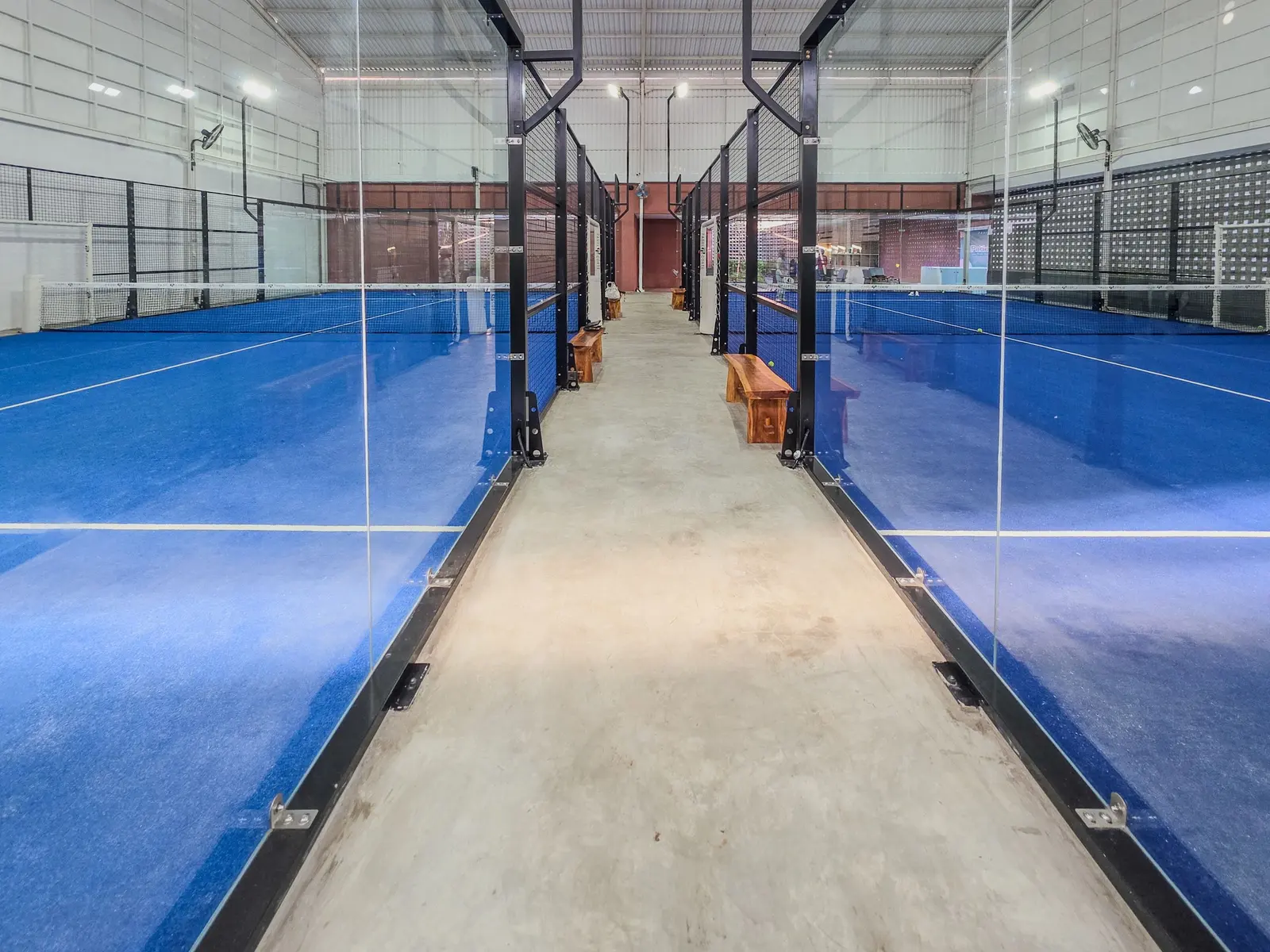 Corridoio tra due campi da padel coperti: vetri temperati a tutta altezza e manto blu ai due lati