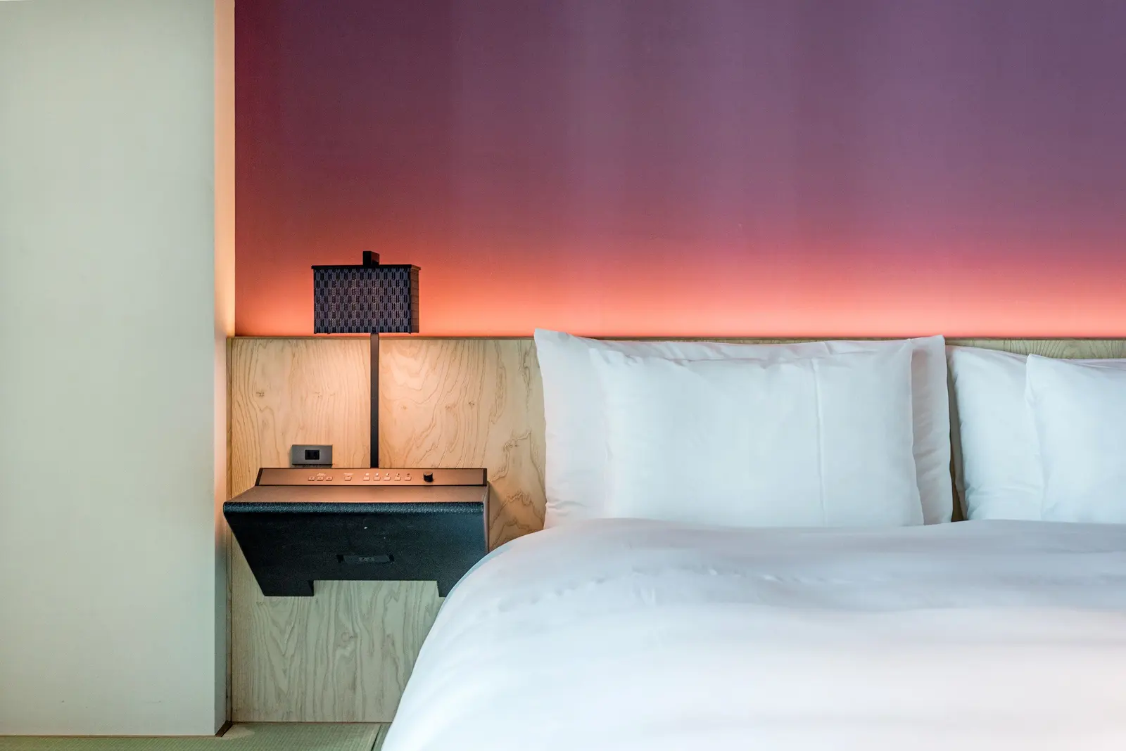 Camera d'albergo: letto bianco, testiera in legno chiaro e parete illuminata dal viola al corallo