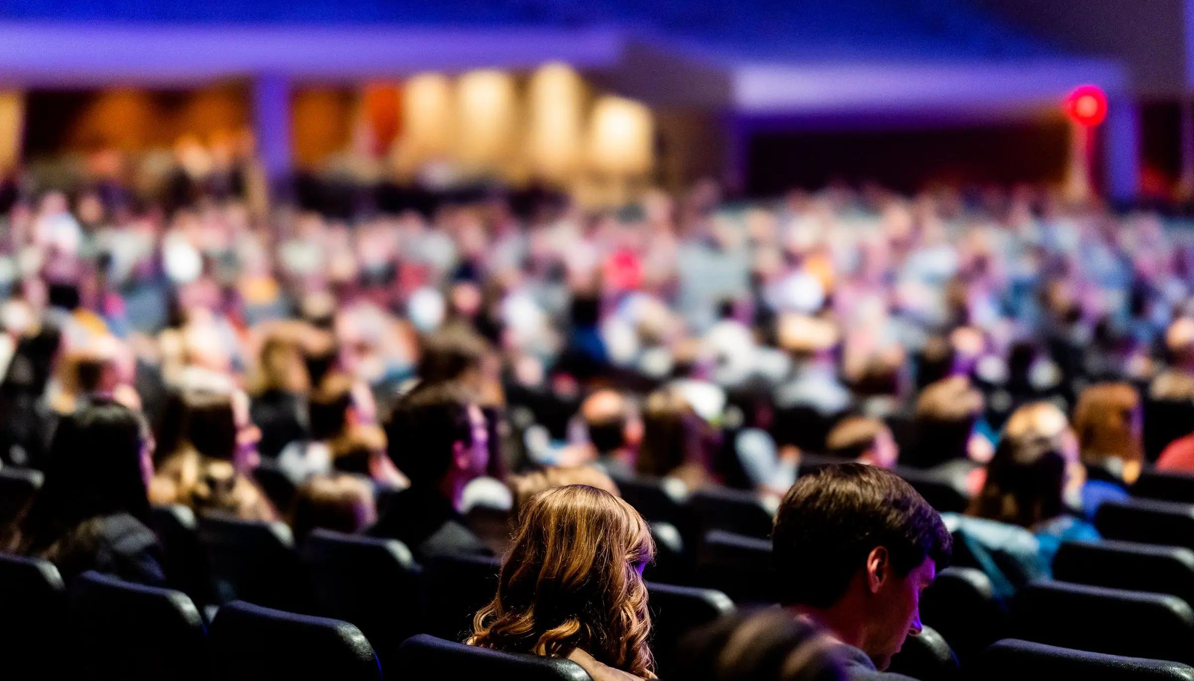 Platea affollata in un auditorium, vista di lato: due persone sedute in primo piano, dietro file di pubblico sfocate sotto luci viola e blu