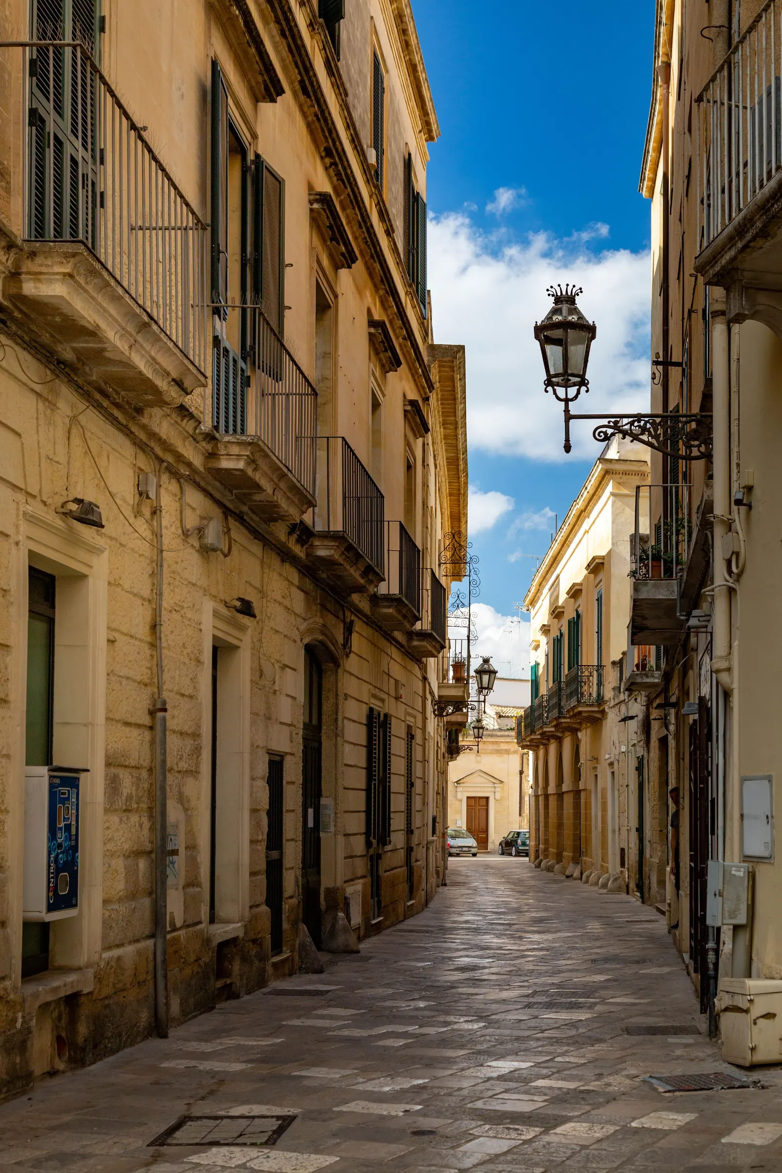 Una via lastricata di un centro storico del Sud, tra palazzi in pietra chiara con balconi in ferro e un lampione a muro