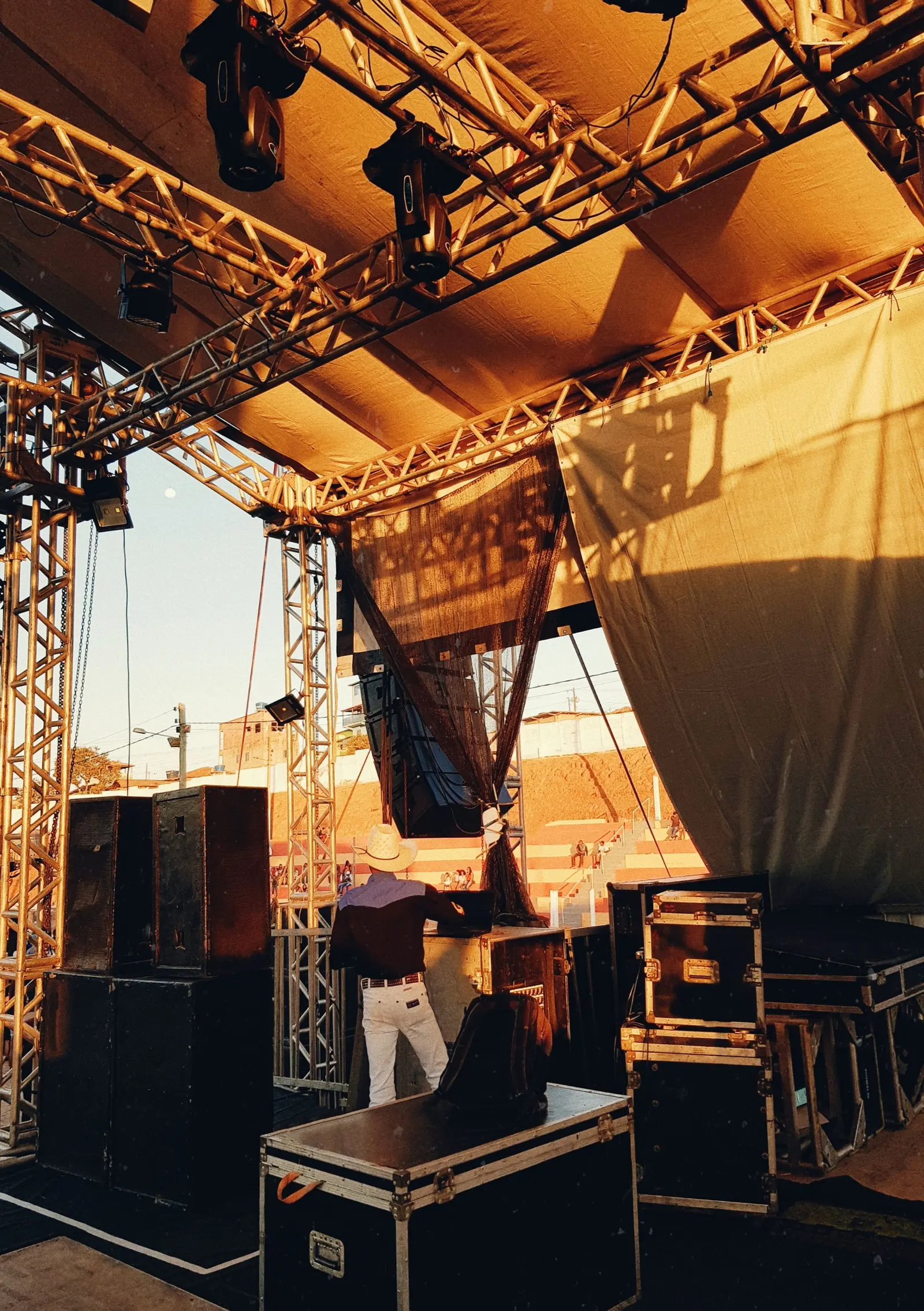 Un palco all'aperto al tramonto, con americane, casse e flight case; un tecnico lavora al mixer