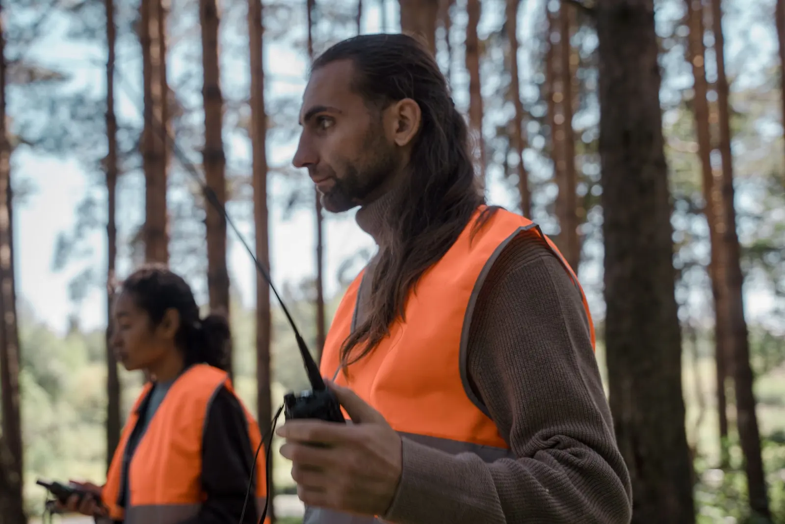 Due persone con il gilet arancione ad alta visibilità in un bosco; lui tiene in mano una radio ricetrasmittente