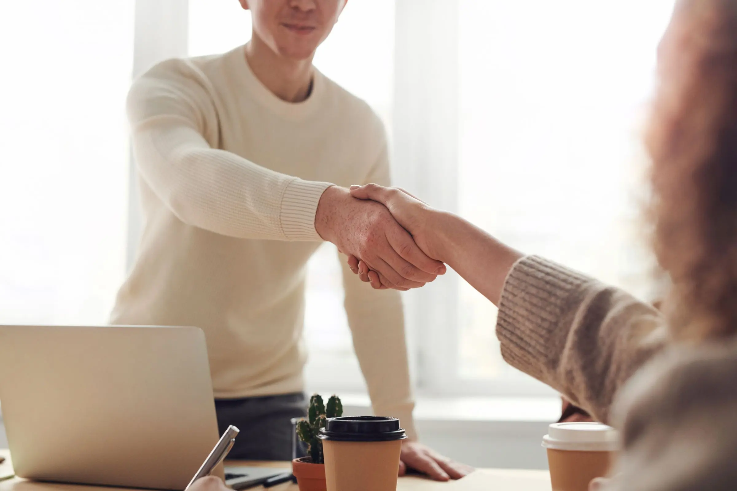Due persone si stringono la mano sopra una scrivania luminosa, con un portatile e due caffè