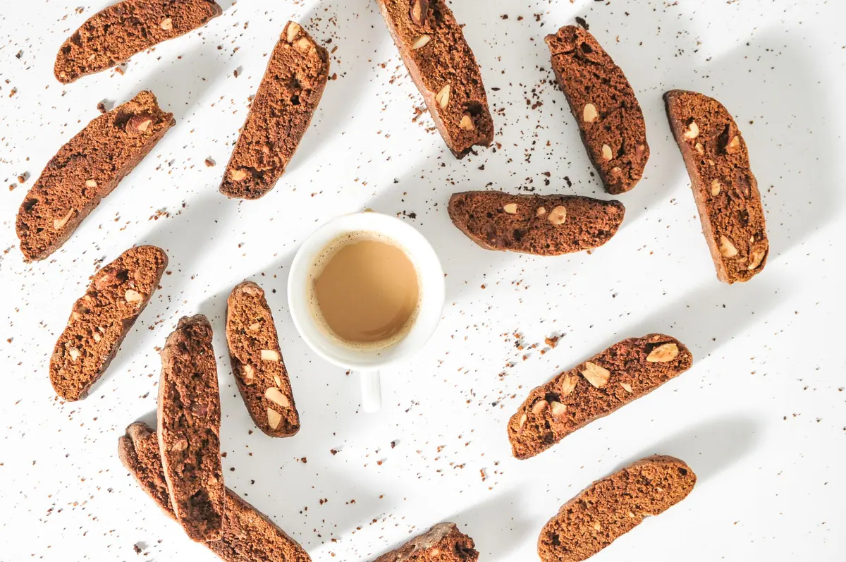 Tozzetti al cacao con mandorle sparsi su fondo bianco accanto a una tazza