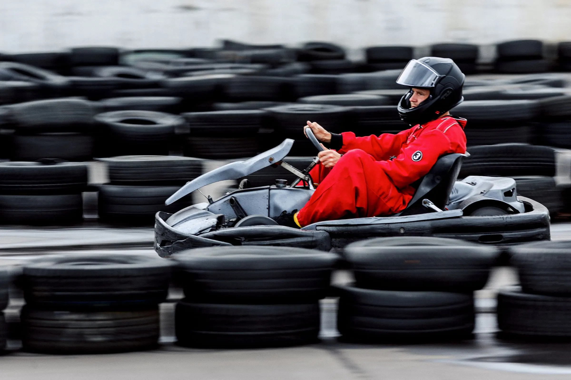 Un pilota in tuta rossa e casco nero su un kart a noleggio, mosso, tra le barriere di gomme