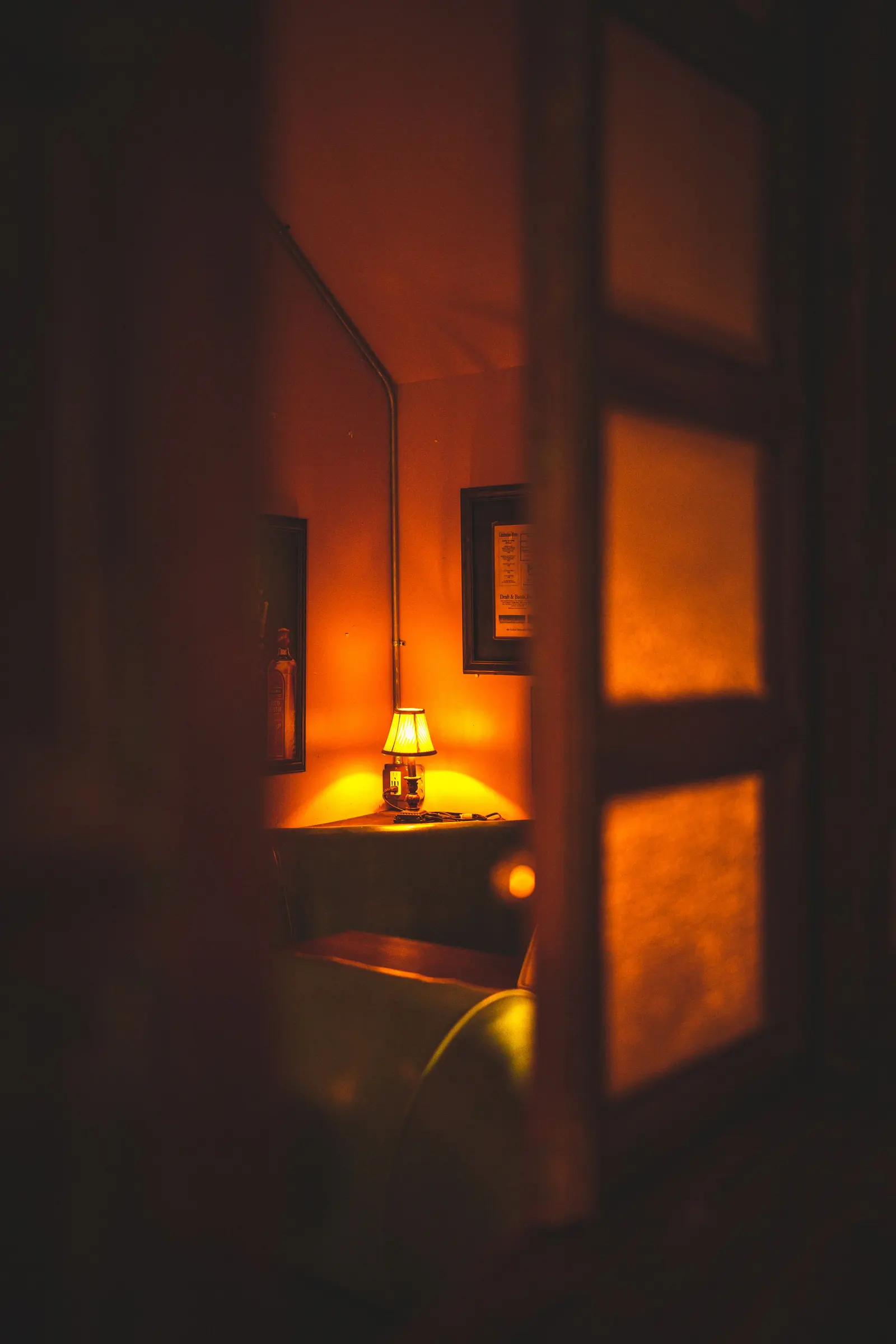 Una lampada accesa su un comodino in una stanza in penombra, vista da una porta socchiusa