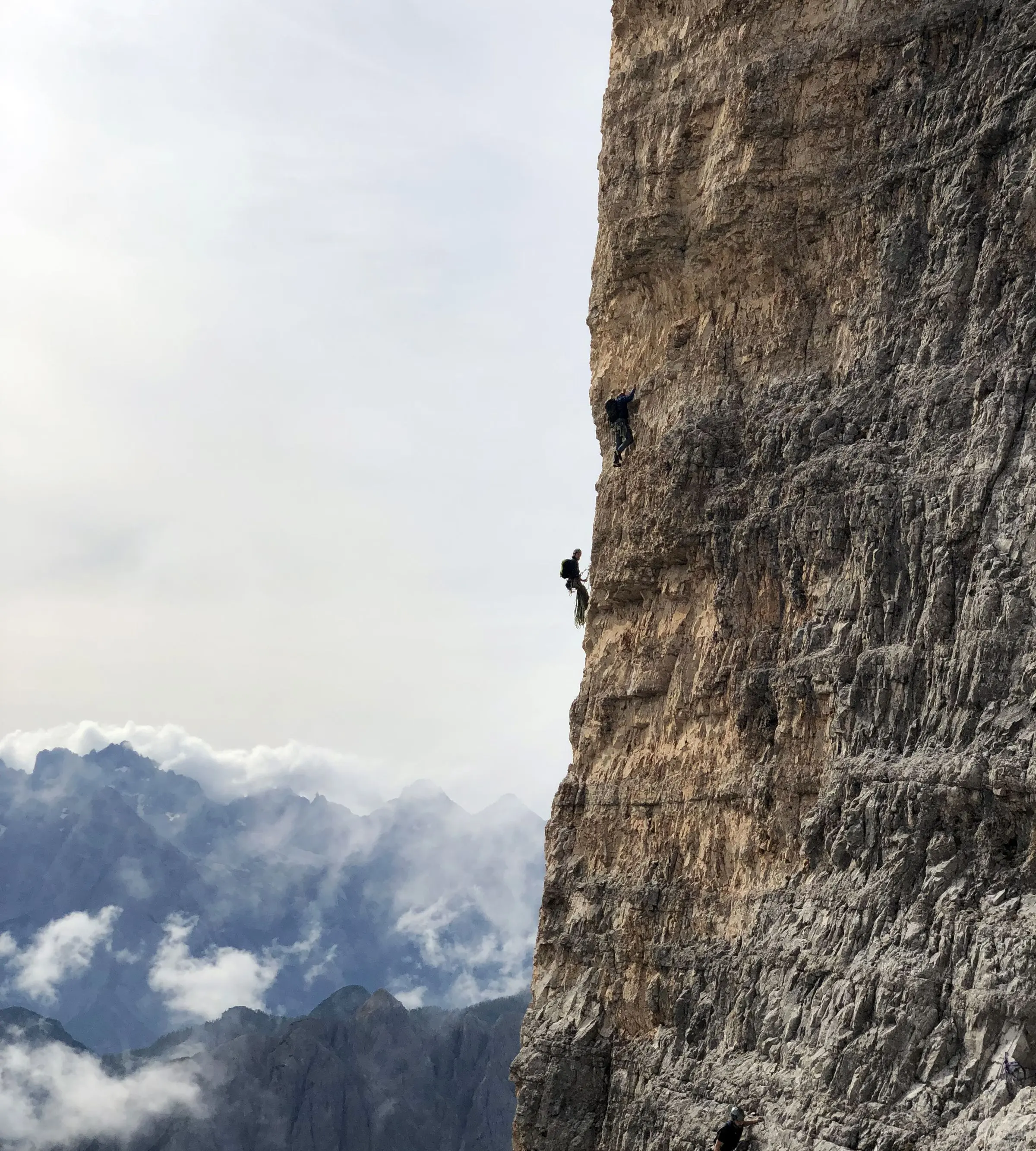 Due alpinisti legati in cordata su una parete calcarea verticale, sullo sfondo cime tra le nuvole