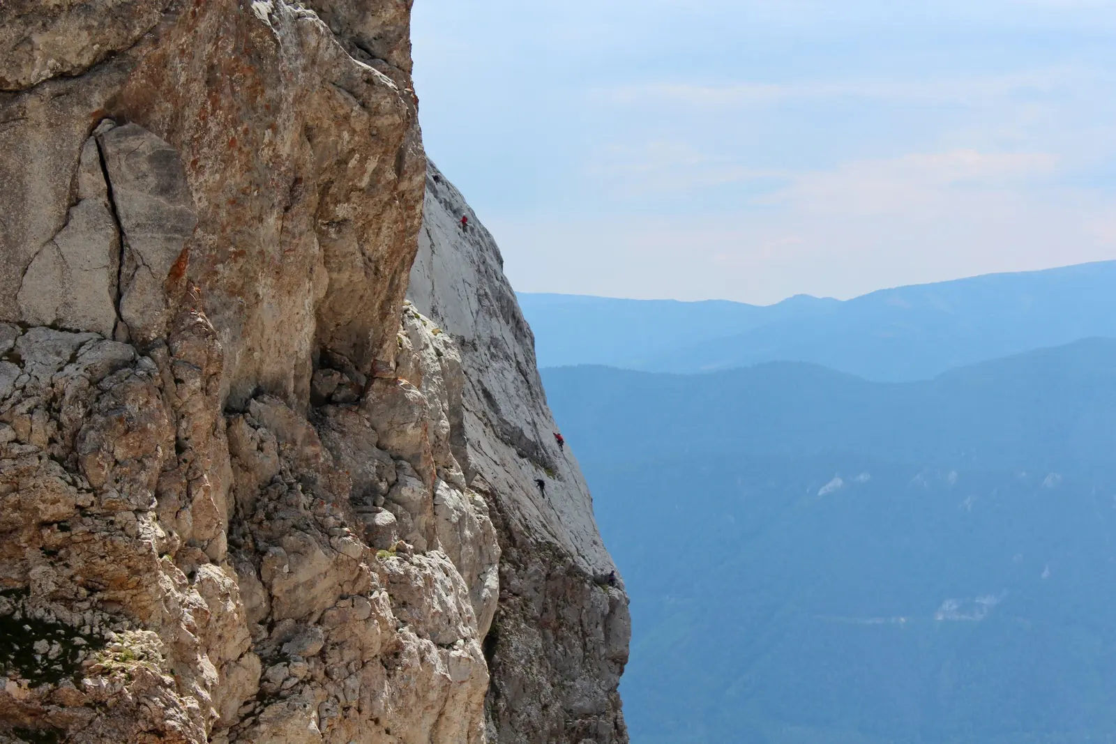 Spigolo di roccia chiara con piccole cordate in salita, dietro una valle prealpina con colline boscose