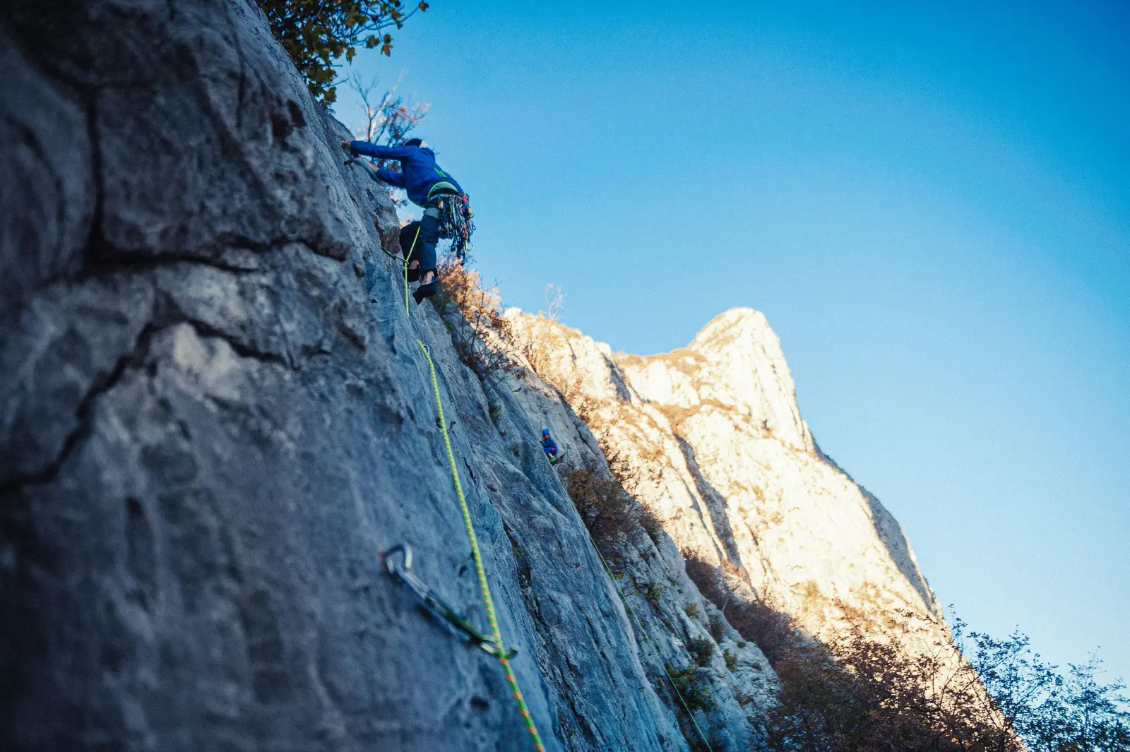 Un arrampicatore sale da primo una placca grigia, la corda passa nei rinvii, cielo blu
