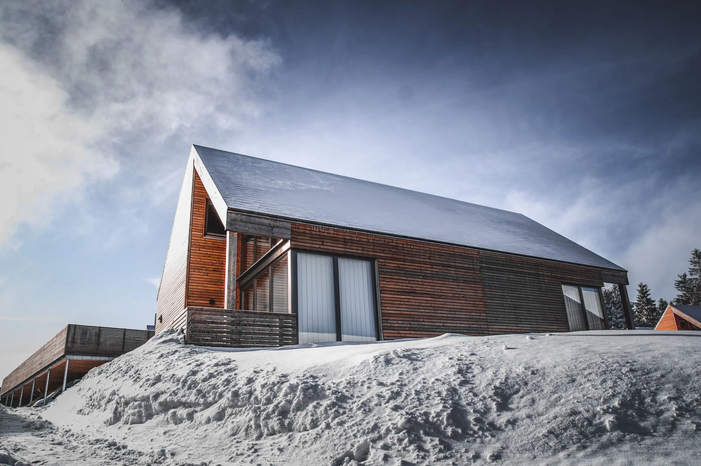 Casa in legno scuro con tetto a falda in lamiera, sulla neve sotto un cielo nuvoloso