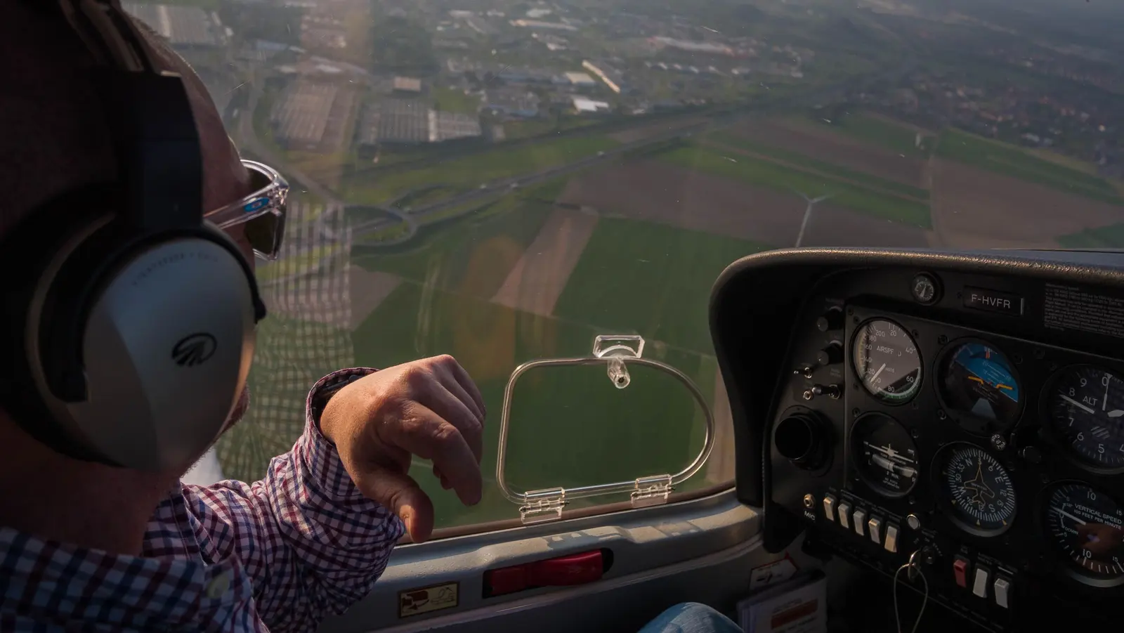 Dalla cabina di un aereo leggero: il pilota con le cuffie, sotto campi e paesi