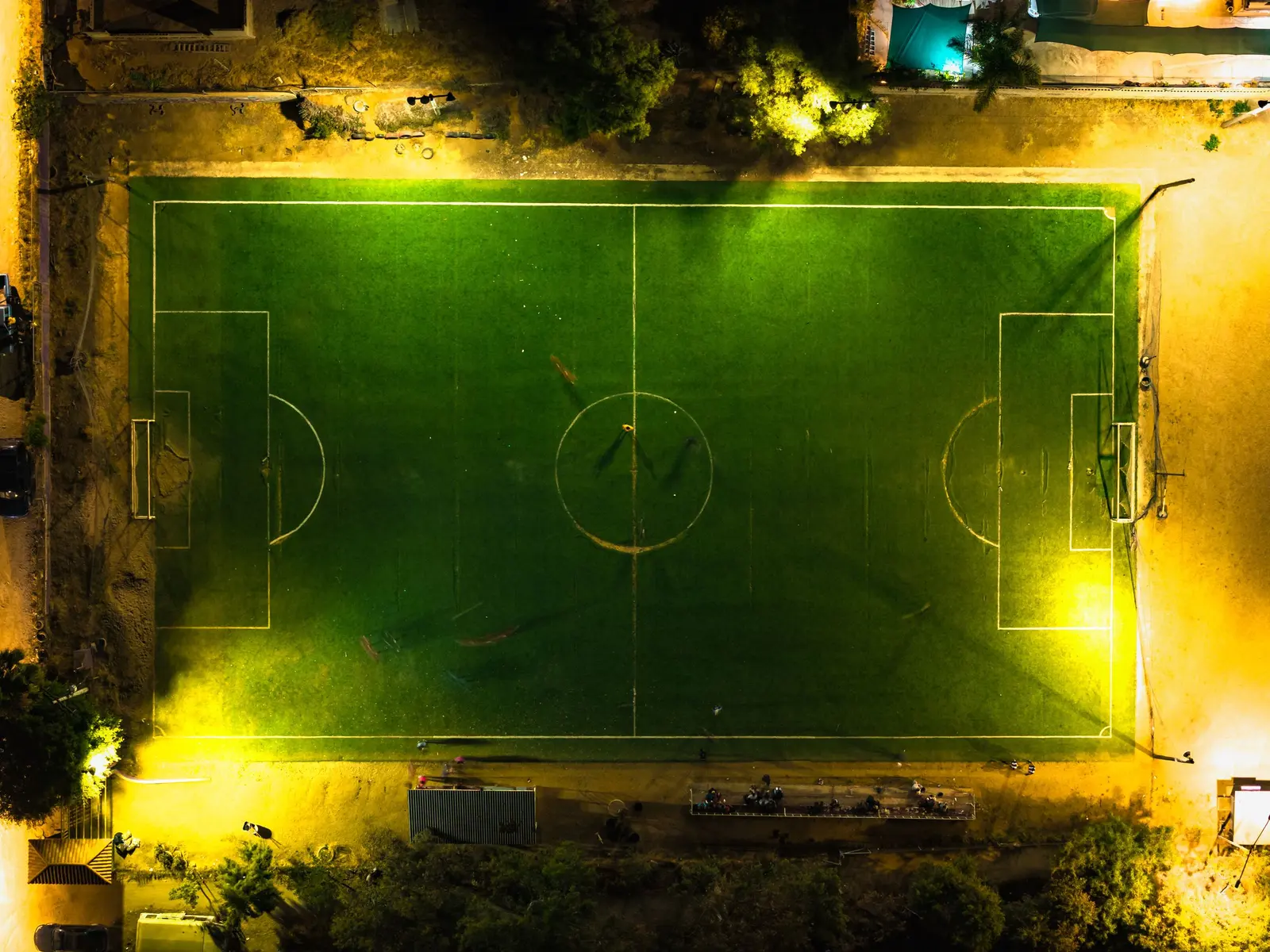 Campo da calcio illuminato di notte visto dall'alto, con le linee bianche e gli alberi intorno