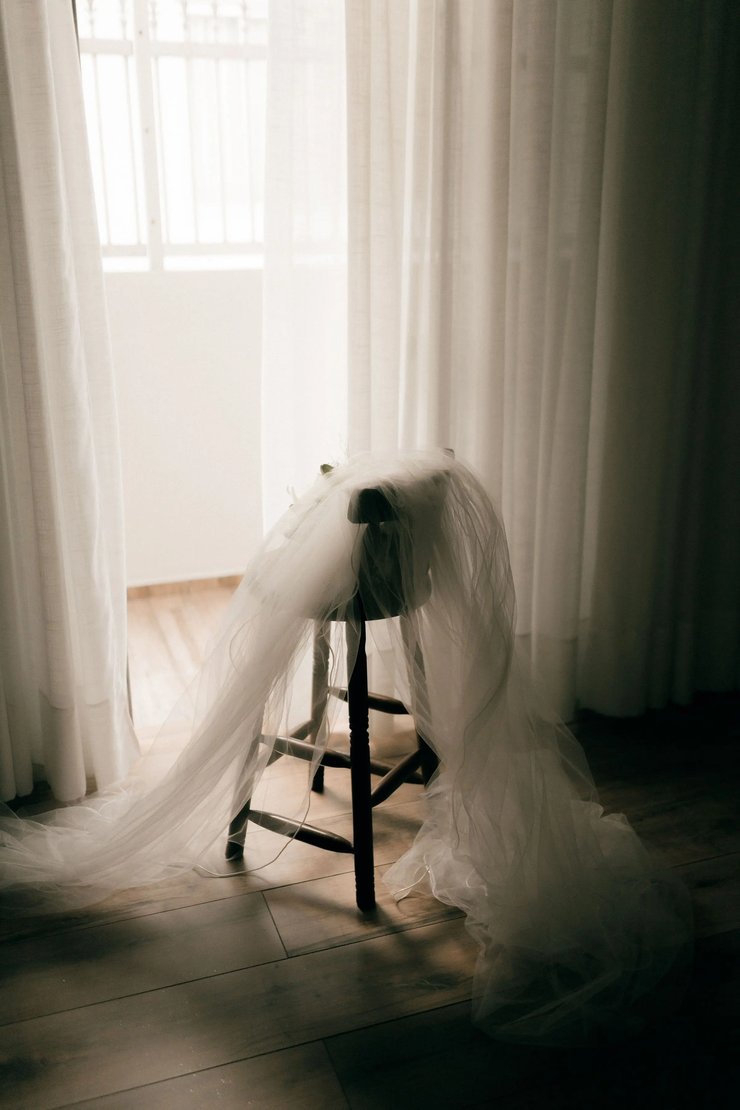 Un velo da sposa appoggiato a uno sgabello, in una stanza con tende bianche