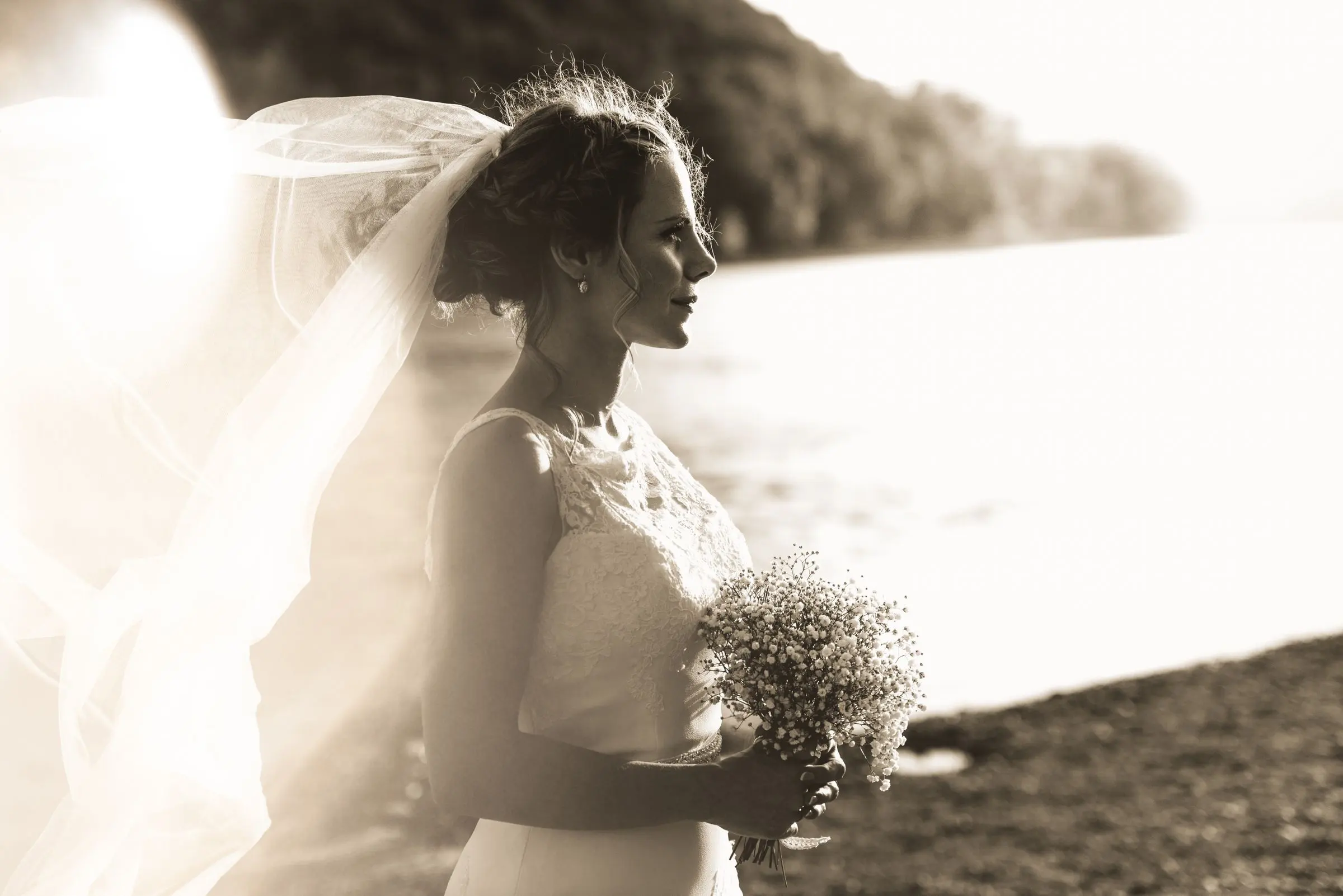 Sposa di profilo in riva al lago con il velo mosso dal vento, toni caldi di seppia