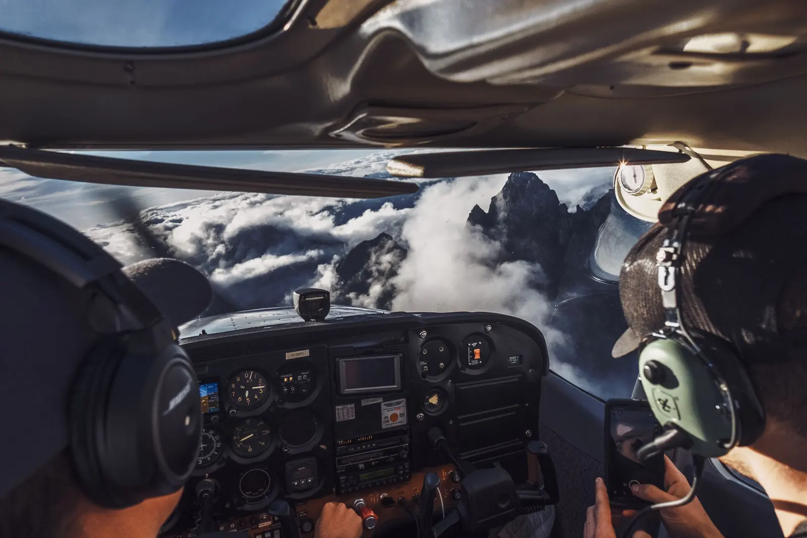 Dalla cabina di un aereo leggero: due piloti con le cuffie e, davanti, cime rocciose tra le nuvole