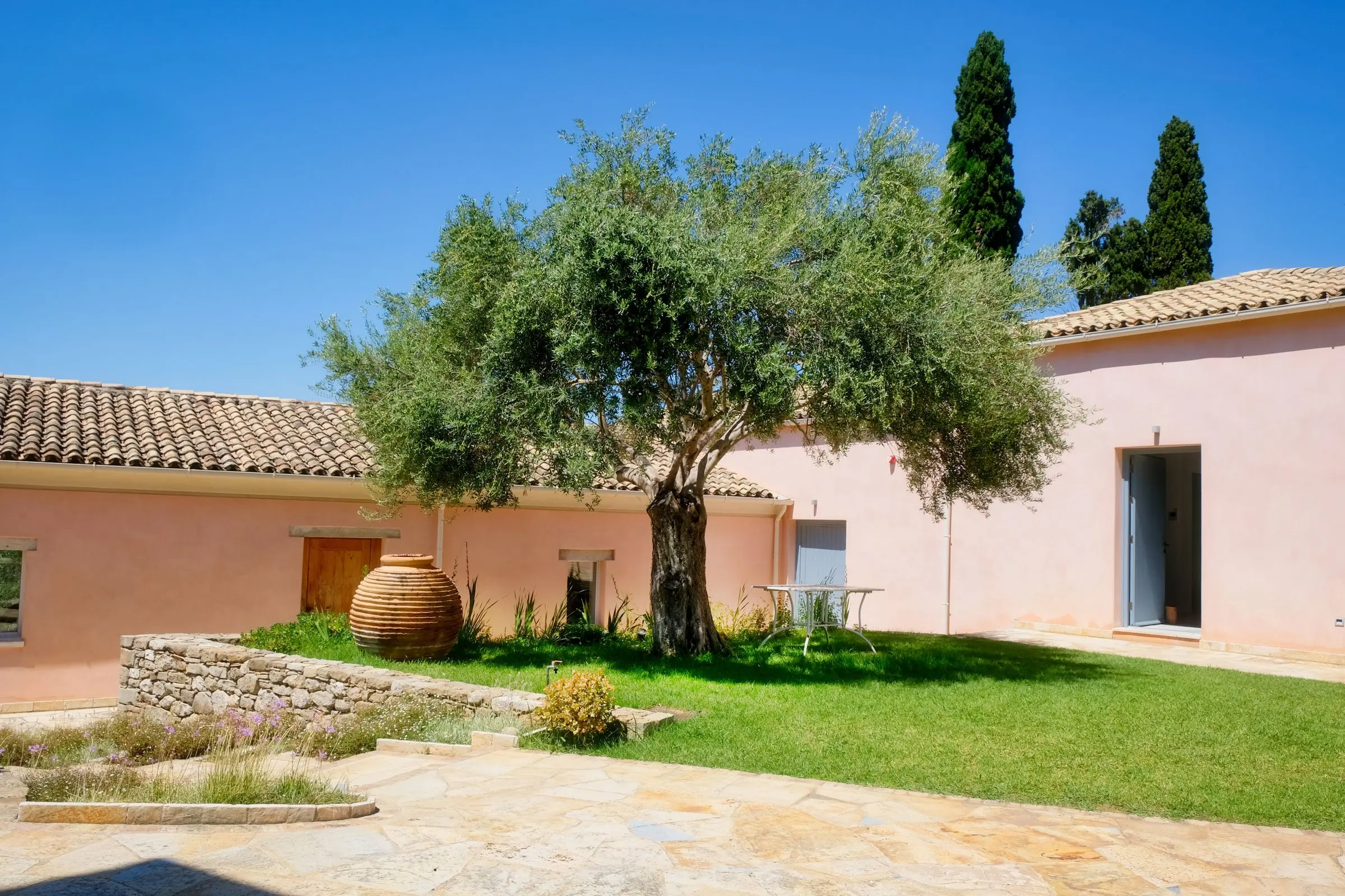 Corte di un casale intonacato rosa, con un grande ulivo su un tappeto di prato, una giara in terracotta e cipressi dietro il tetto